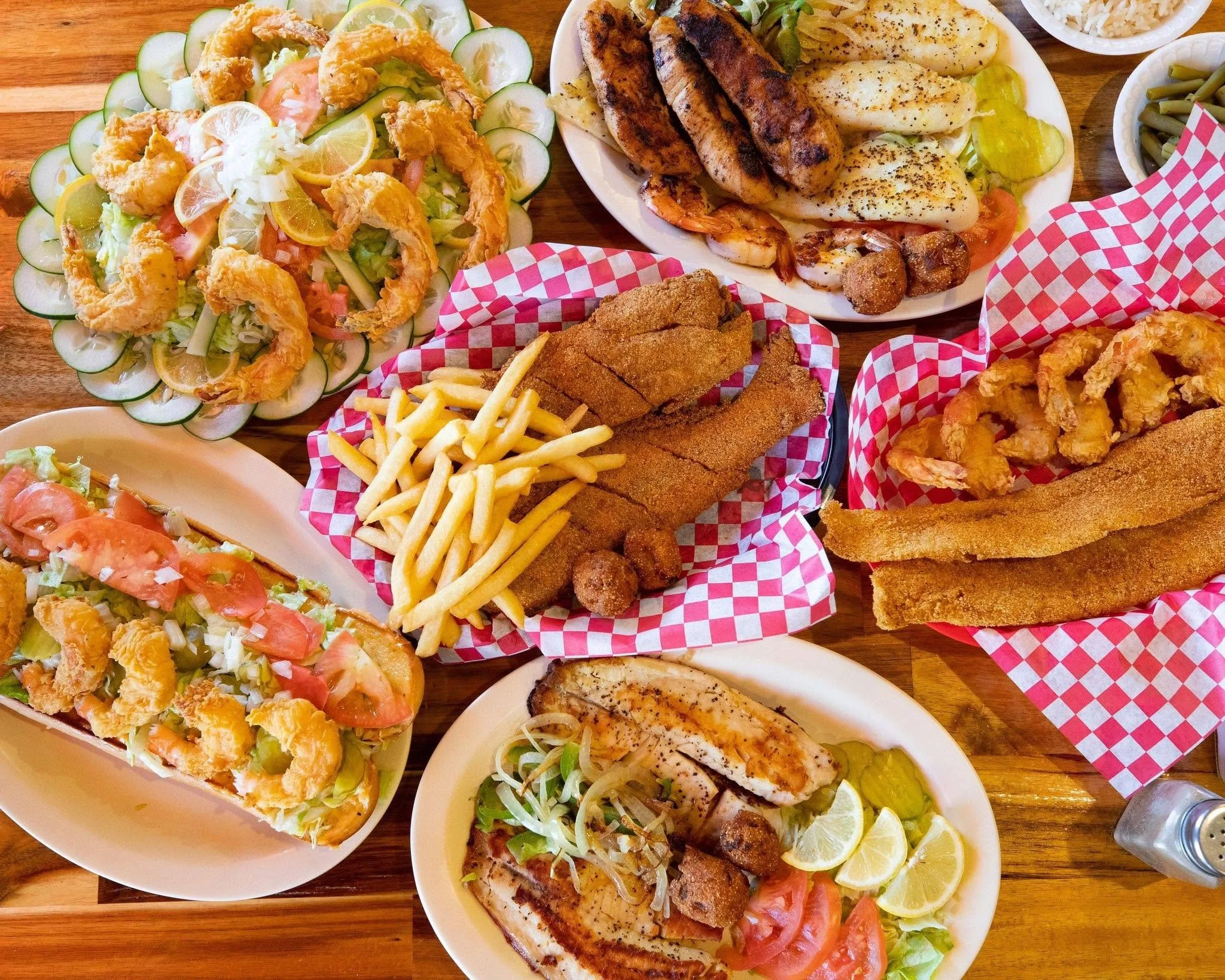 Various plates of seafood dishes including fried shrimp, grilled fish, fried fish fillets, and shrimp with lemon wedges, along with sides of French fries, salad, and vegetables on a wooden table.