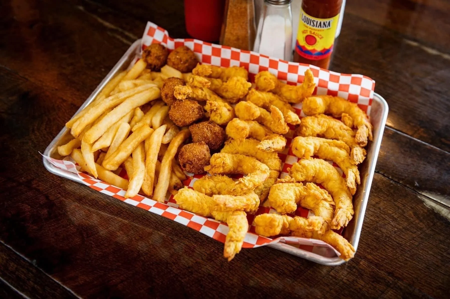 A basket of French fries, chicken nuggets, and fried shrimp. The basket is lined with red and white checkered paper, on a dark wooden table, with salt, pepper, and hot sauce bottles in the background.