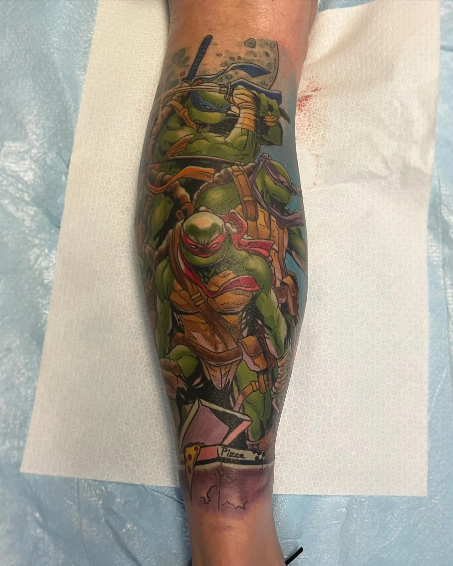 Colorful tattoo of the Teenage Mutant Ninja Turtles on a person’s leg, featuring four turtles in combat gear, with a pizza slice at the bottom by Jesse Smith at Human Production Gallery in Bethel Park, PA.