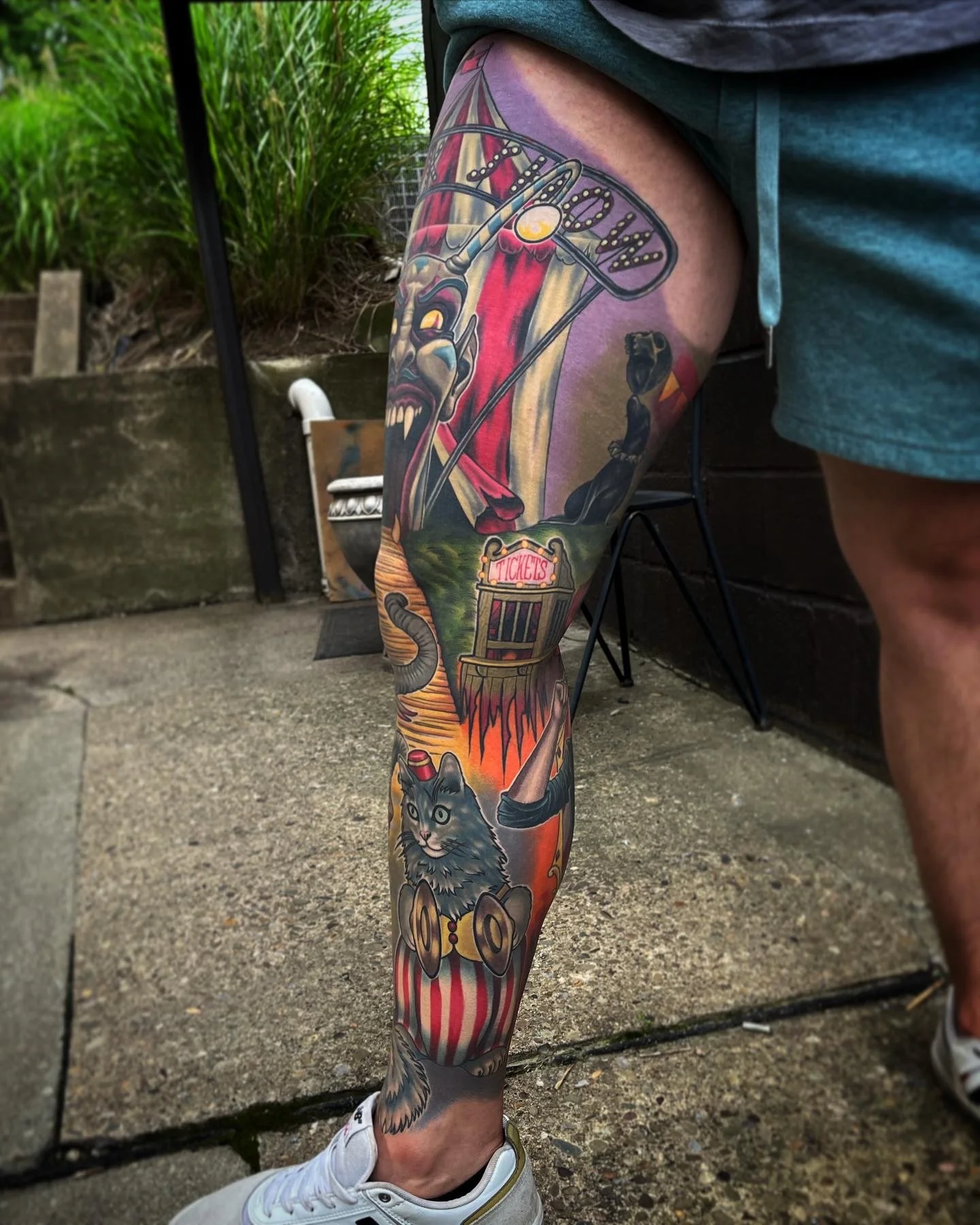Award-winning color tattoo sleeve on person's leg featuring a cat, a ticket booth, and various vibrant elements by Jesse Smith serving Bethel Park, PA.