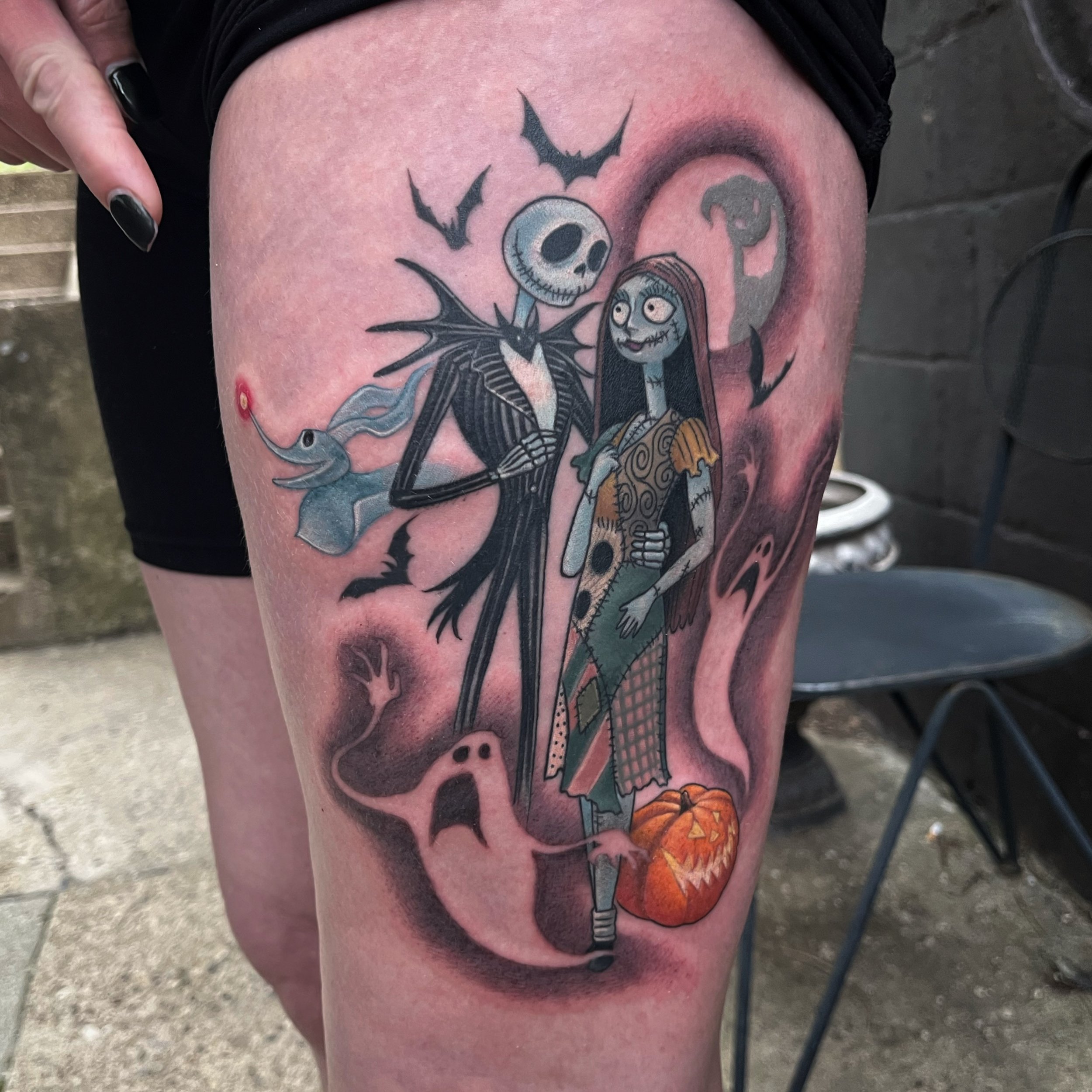 Tattoo on person's thigh depicting Jack Skellington, Sally, a bat, a ghost, and a pumpkin in a cartoon style by Jesse Smith at Human Production Gallery in Bethel Park, PA.