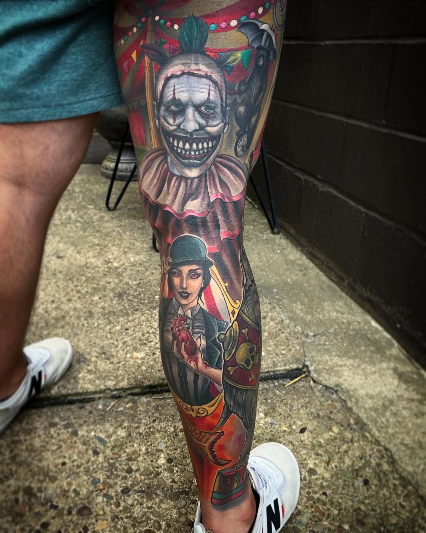 A person's leg with a detailed tattoo featuring a clown with a sinister smile and circus-themed elements by Jesse Smith in Pittsburgh, PA.