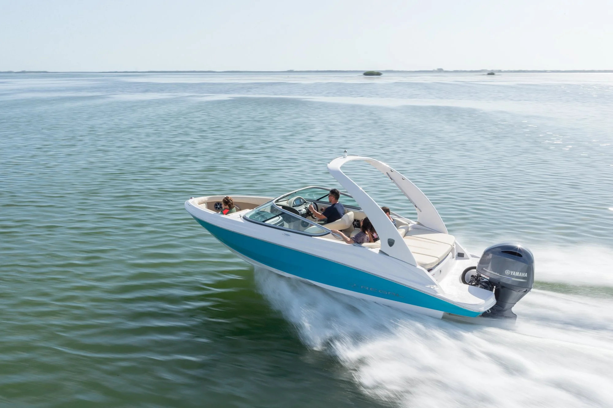 A white and blue motorboat with four people on board speeding across a calm body of water. The boat has a Yamaha outboard motor and a sleek, modern design. The water is greenish and the sky is clear with a few distant trees visible on the horizon.