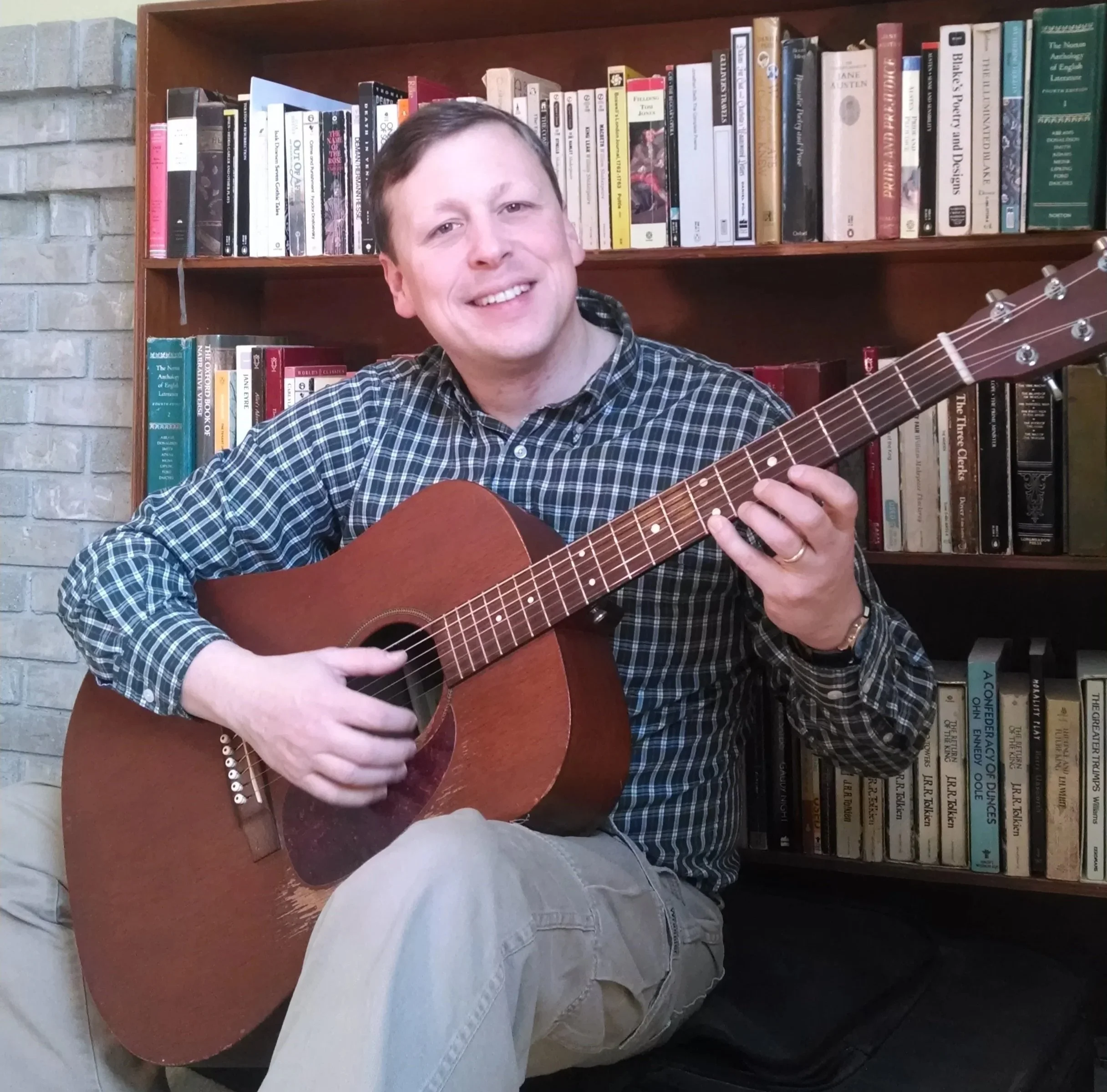 Michael Straight holding his acoustic guitar sitting in a guitar studio with wooden bookshelves filled with books in the background.