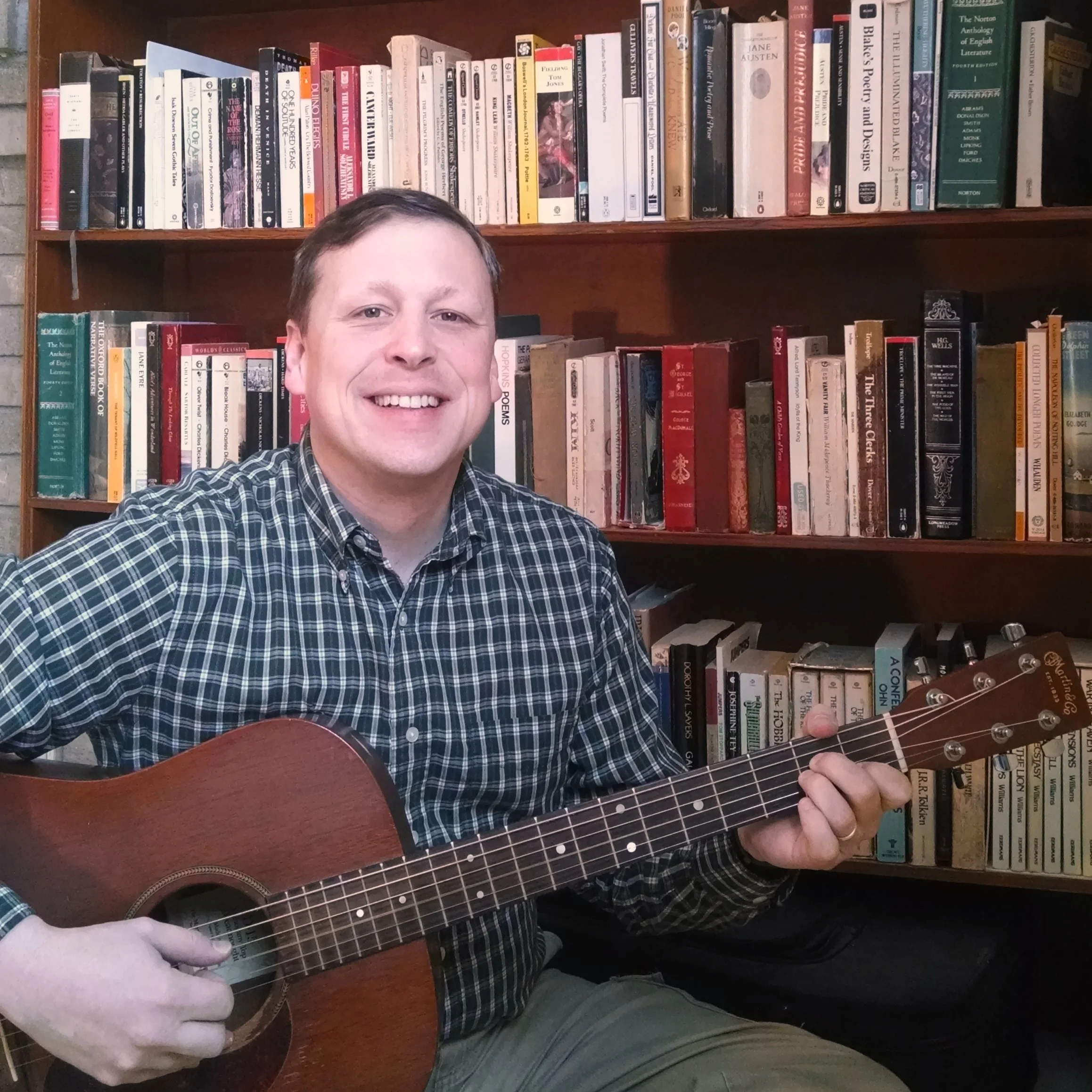 A man holding an acoustic guitar in a room with wooden bookshelves filled with books in the background.