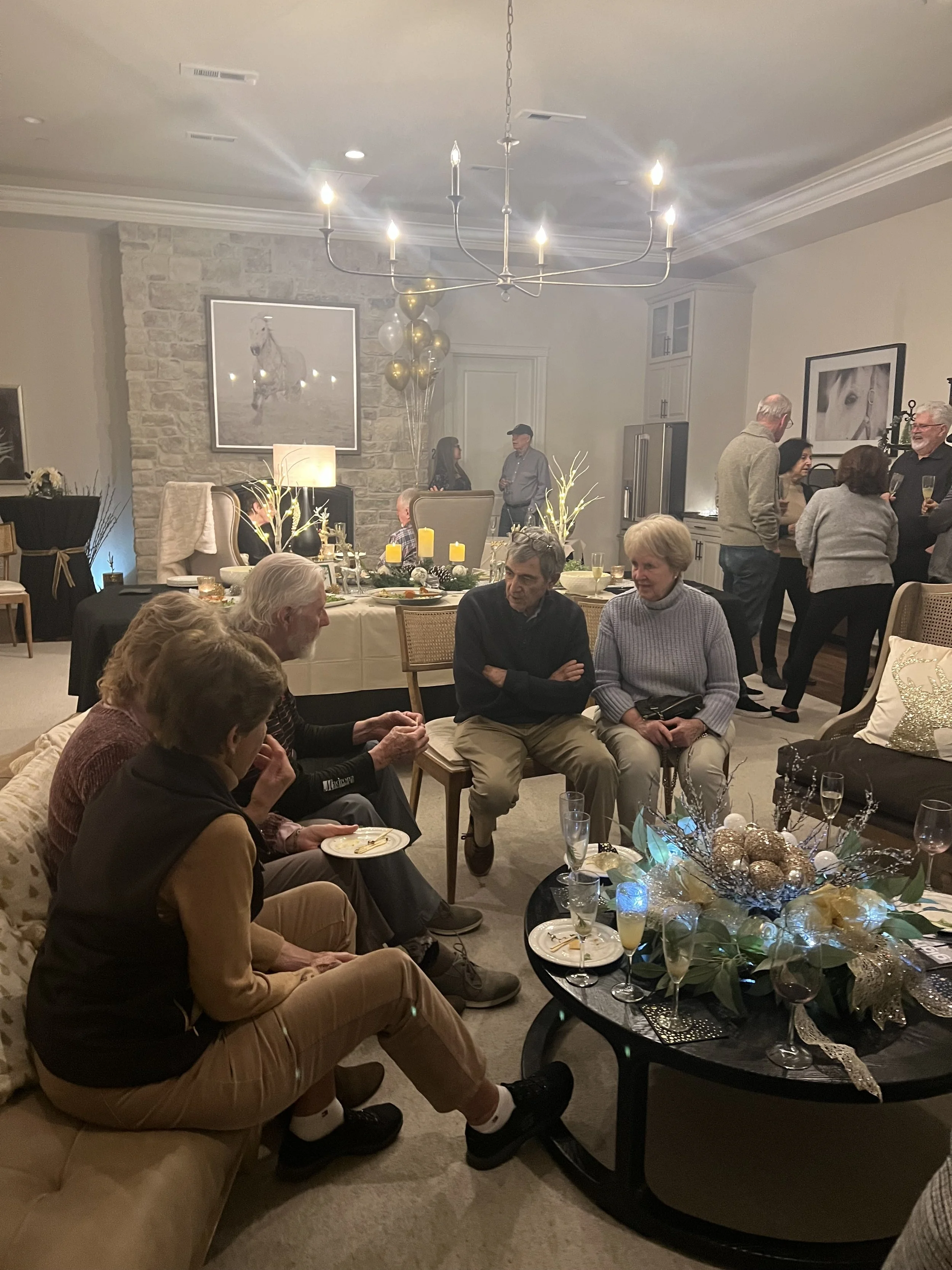 A group of people gathered at a holiday party in a living room decorated with festive decorations and a chandelier overhead. Some are seated, others are standing and talking, with food and drinks on the table.