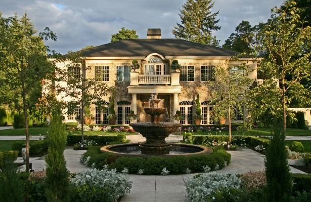 A large, elegant mansion with a fountain in front, surrounded by landscaped gardens and trees.