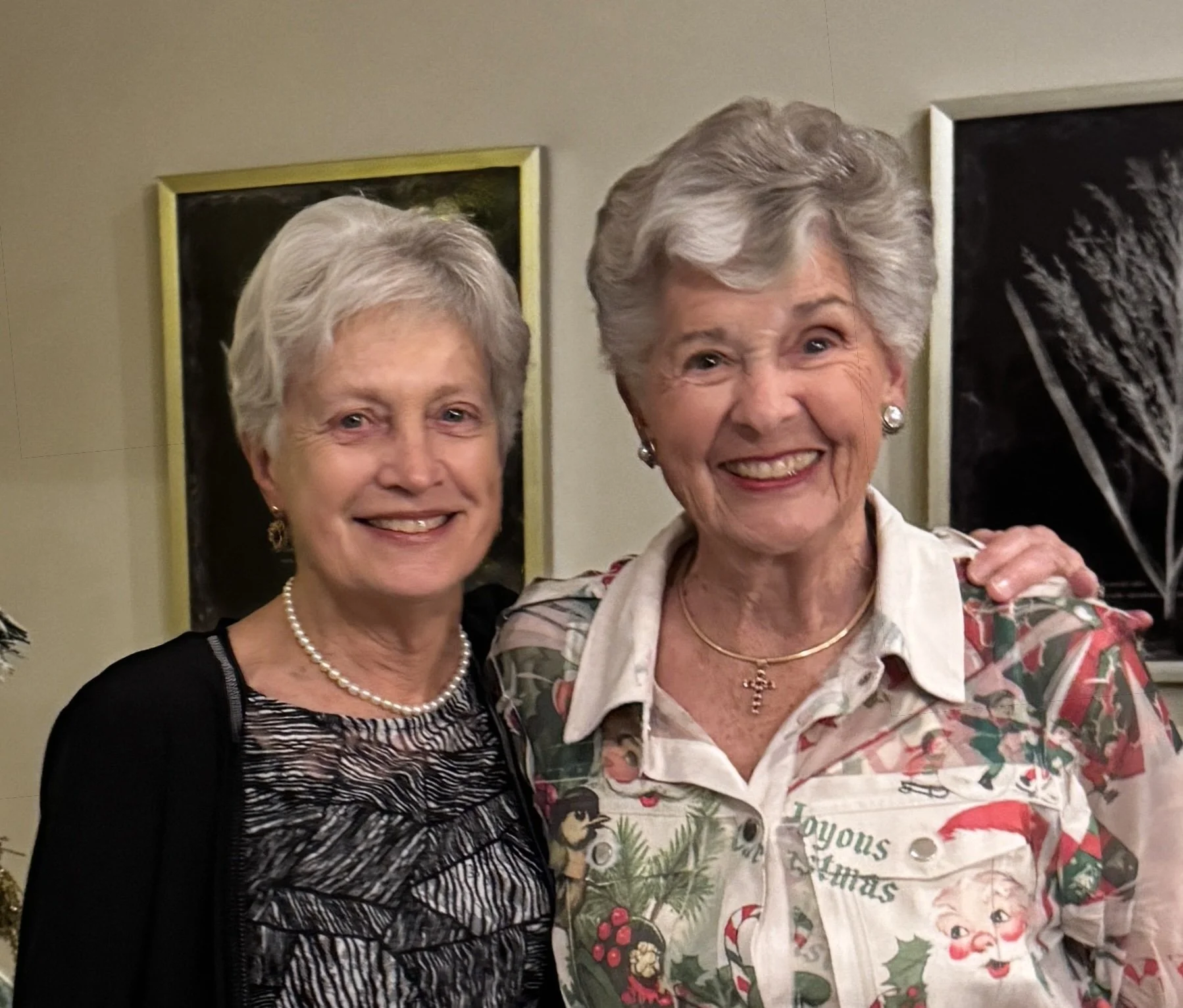 Two women are smiling and posing for a photo together indoors, standing in front of framed art on a beige wall. The woman on the left is wearing a black and white patterned top, pearl necklace, and earrings. The woman on the right is wearing a festive holiday shirt with Christmas-themed illustrations, jewelry, and has short gray hair.