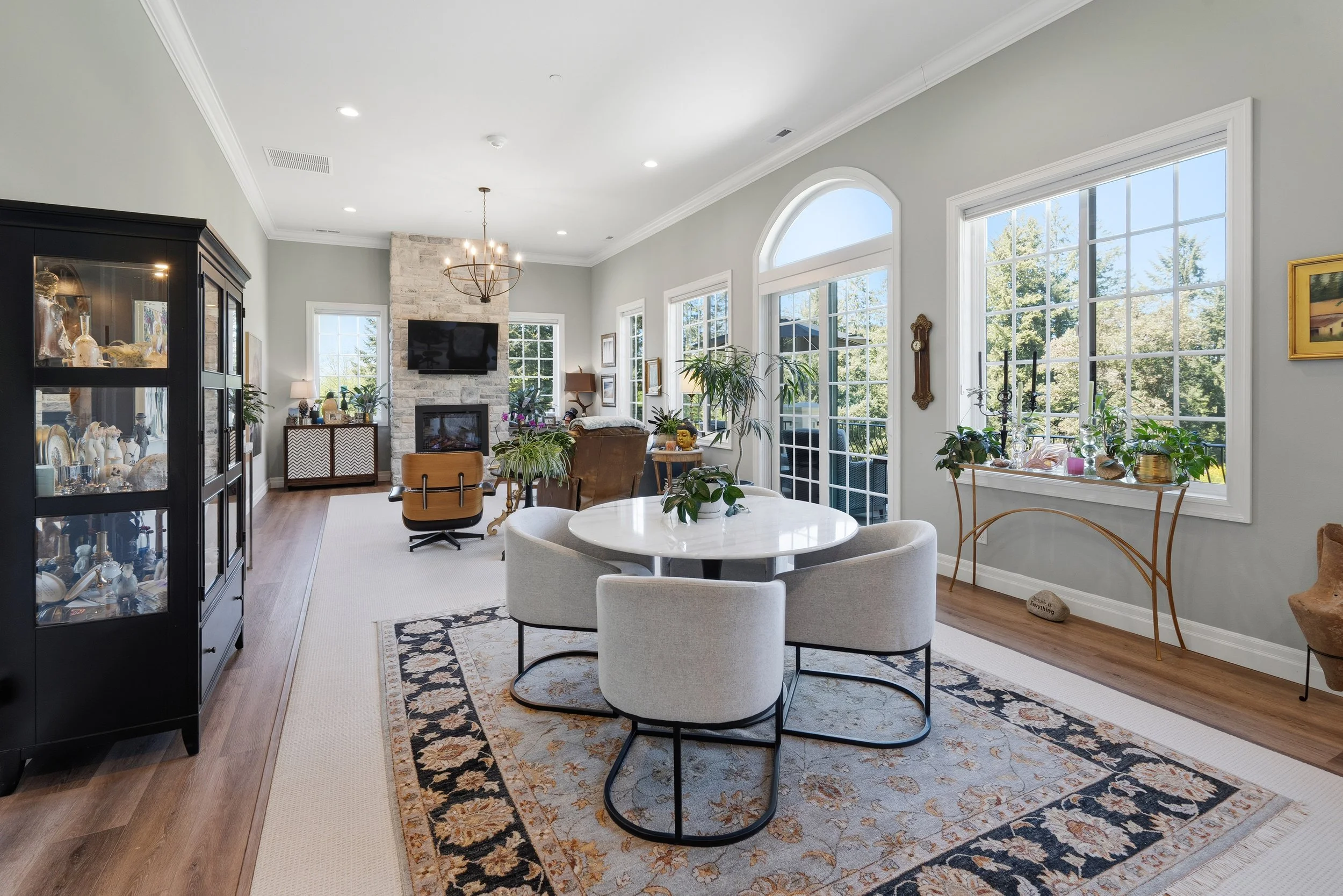 Bright living room with large windows, a stone fireplace, modern chandelier, black display cabinet, plants, and various decorations.