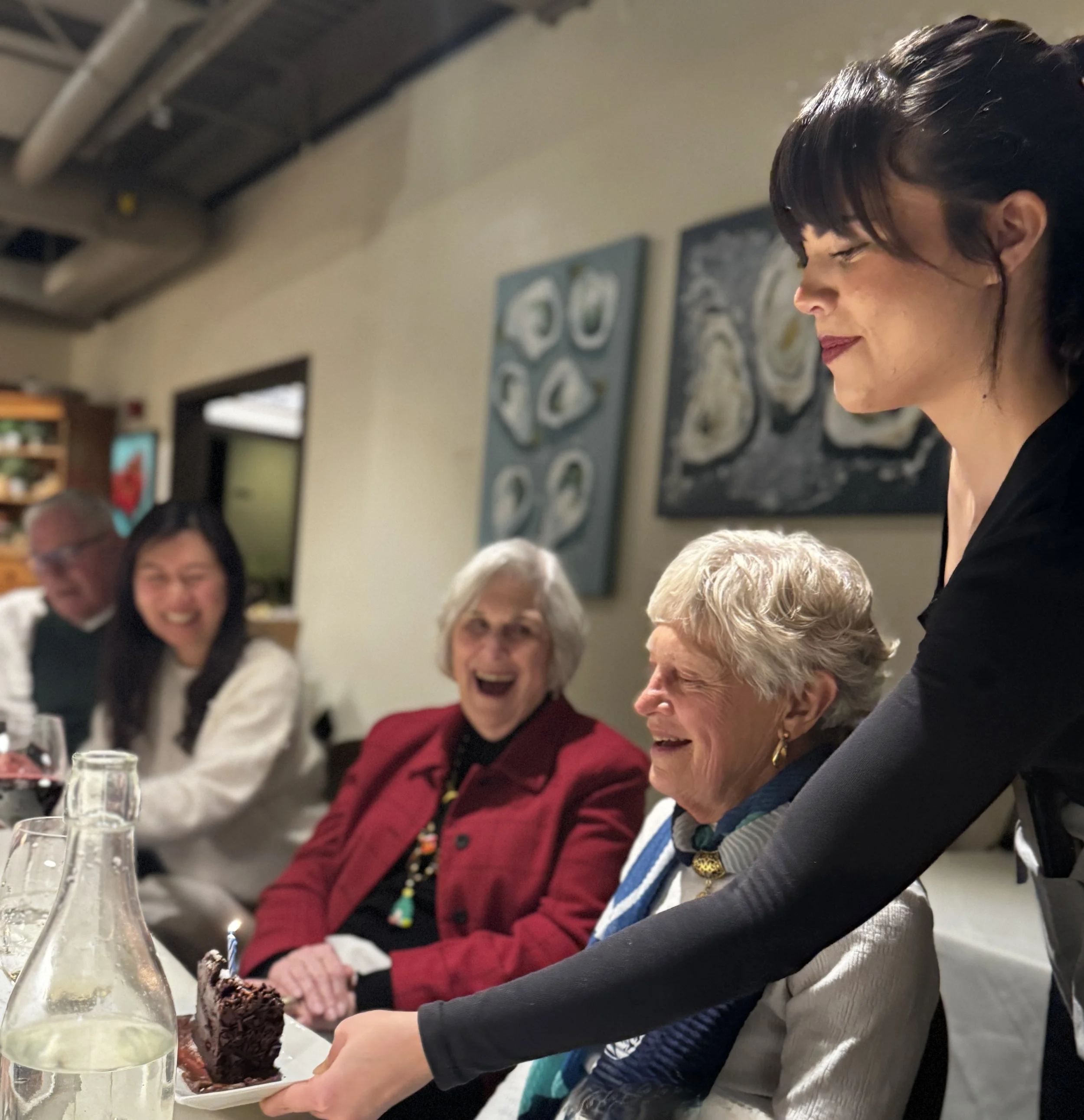 People at a dining table, with a woman serving a slice of chocolate cake, glasses of wine, and a water pitcher in the foreground, and several smiling individuals in the background.