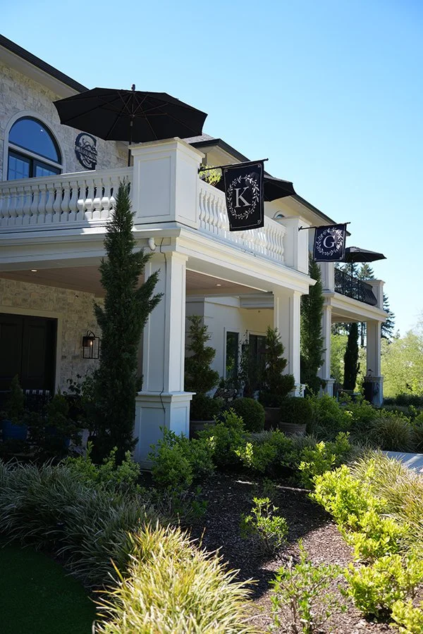 Exterior view of a multi-story house with a white porch, black umbrellas, and hanging banners displaying the letters K and G, surrounded by green plants and trees under a clear blue sky.