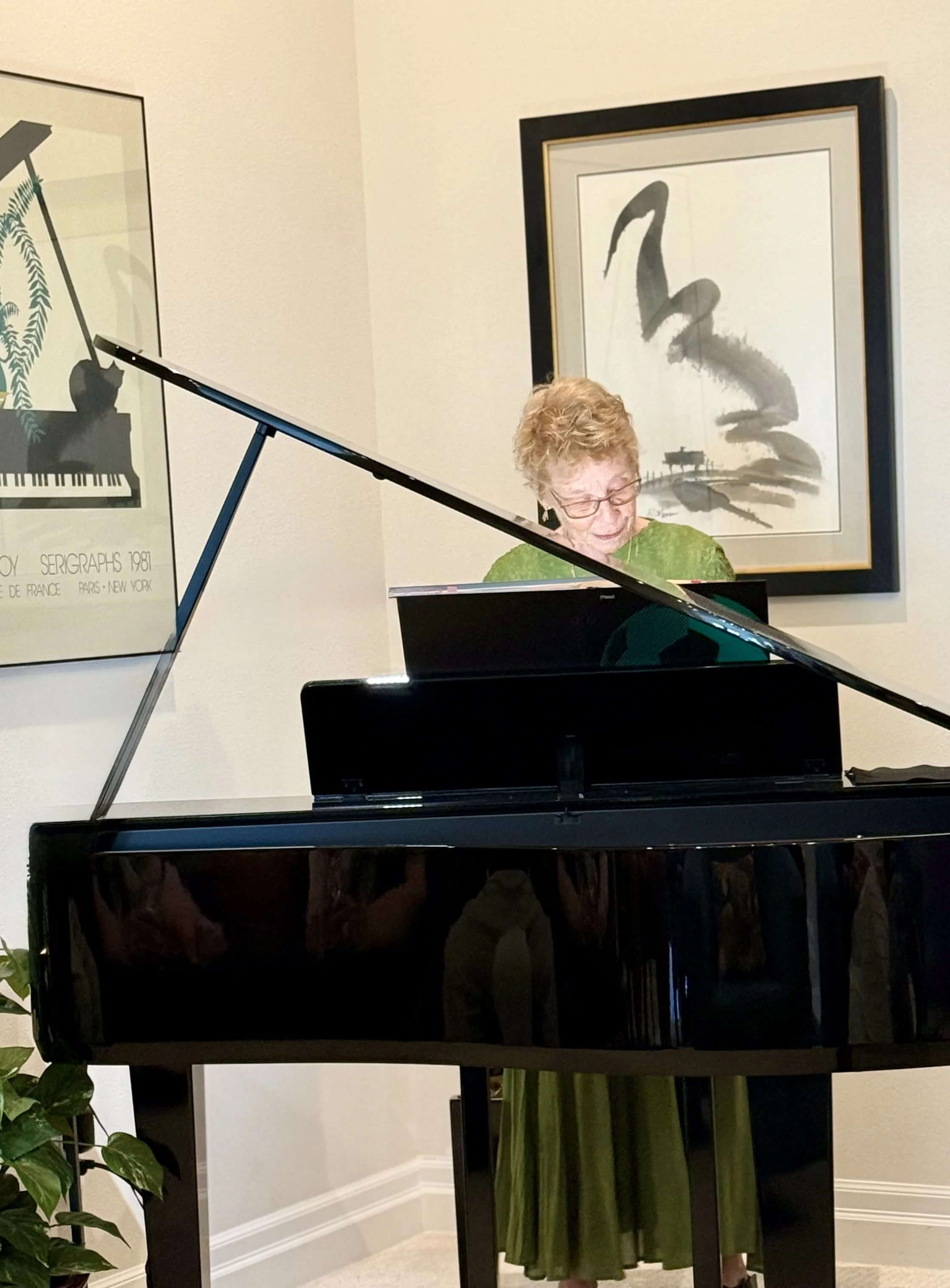 A woman playing a black grand piano in a room with framed artwork on the wall, a green curtain below the table, and a potted plant in the corner.