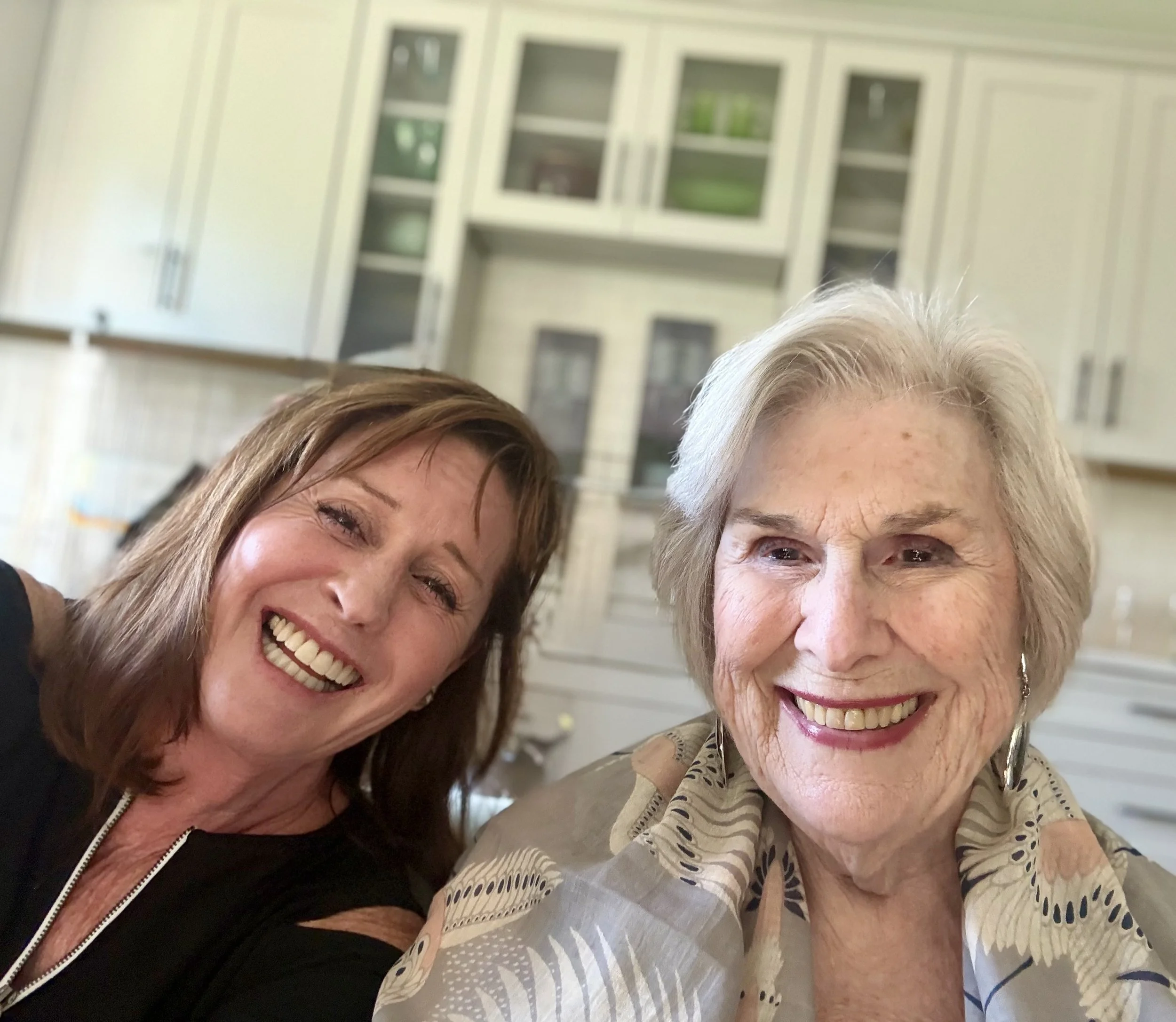 Close-up of two smiling women, one with brown hair and the other with white hair, in a home setting with cabinets in the background.