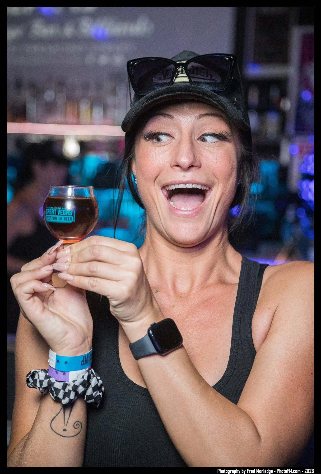 A woman holding a small glass of beer at an indoor event with colorful lights and a blurred background. She has a black cap with sunglasses on top, a black tank top, a smartwatch, and colorful wristbands. She has a tattoo of a whimsical face on her r