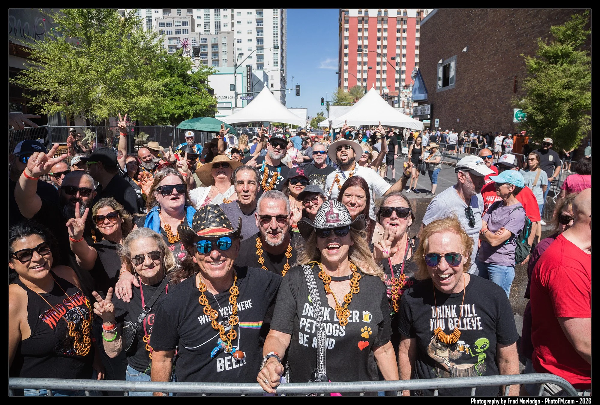 A large group of people gathering outdoors in a city street during daytime, many wearing Black Lives Matter T-shirts, orange necklaces, and sunglasses, with tents, trees, and tall buildings in the background.