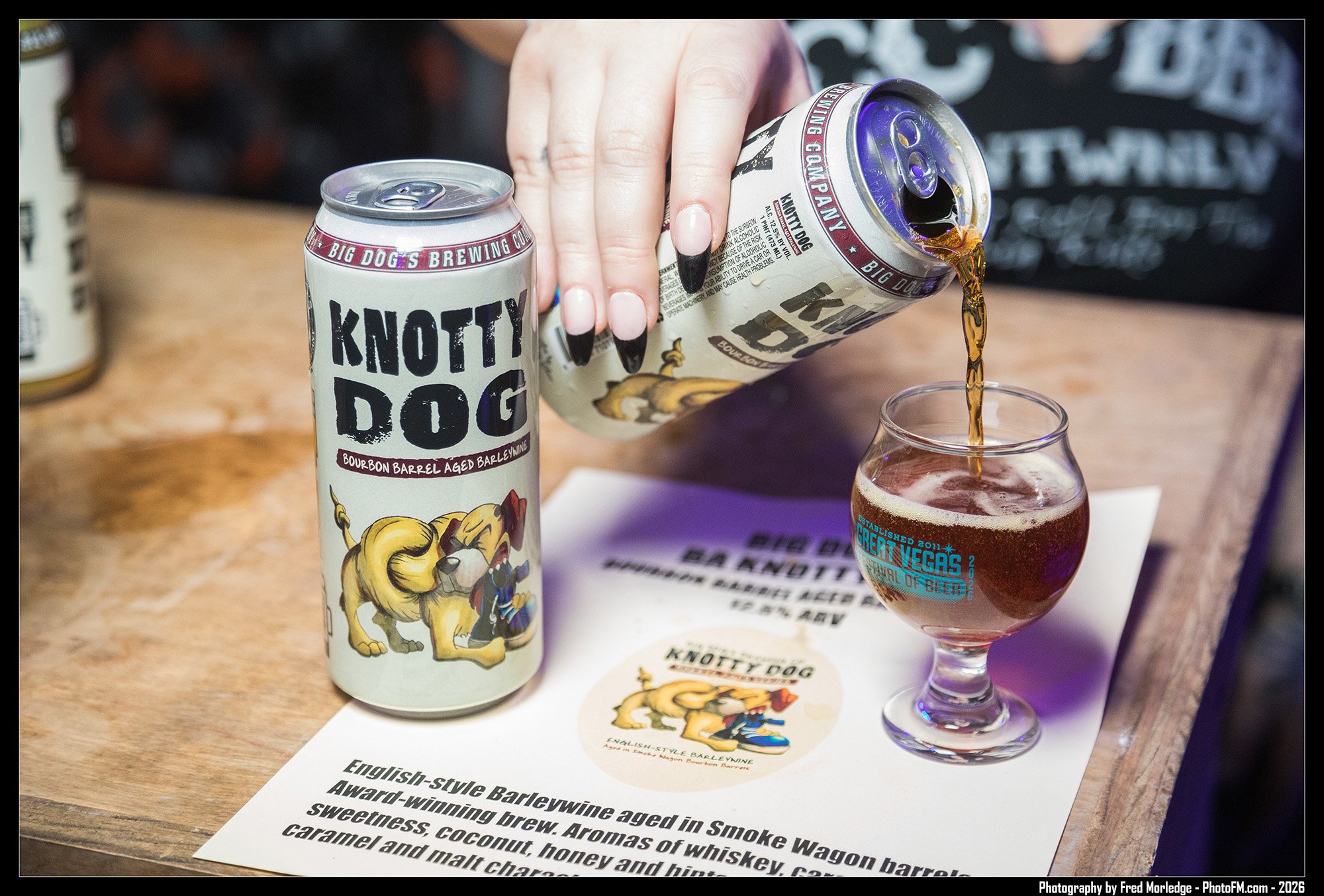 A person pouring beer from a can labeled 'Knottty Dog' into a glass. The can features cartoon dog characters. A printed description of the beer is on the table.