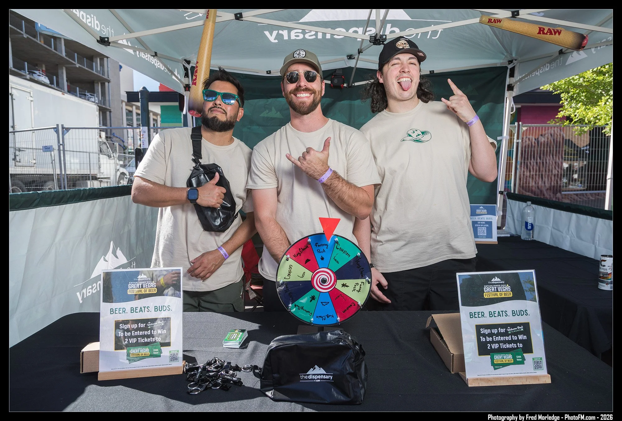 Three men standing behind a table at a festival booth with a spinning prize wheel. The booth displays signs indicating a raffle for VIP tickets, with promotional items like sunglasses, keychains, and wristbands visible.