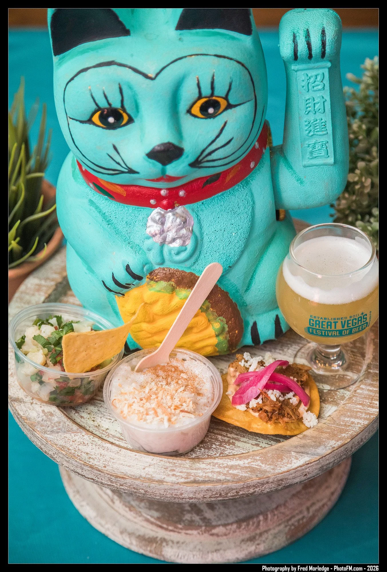 A teal-colored Maneki-neko cat statue with black and yellow eyes, a red collar, and Japanese characters on its raised paw, surrounded by small bowls of food and drinks on a round tray.