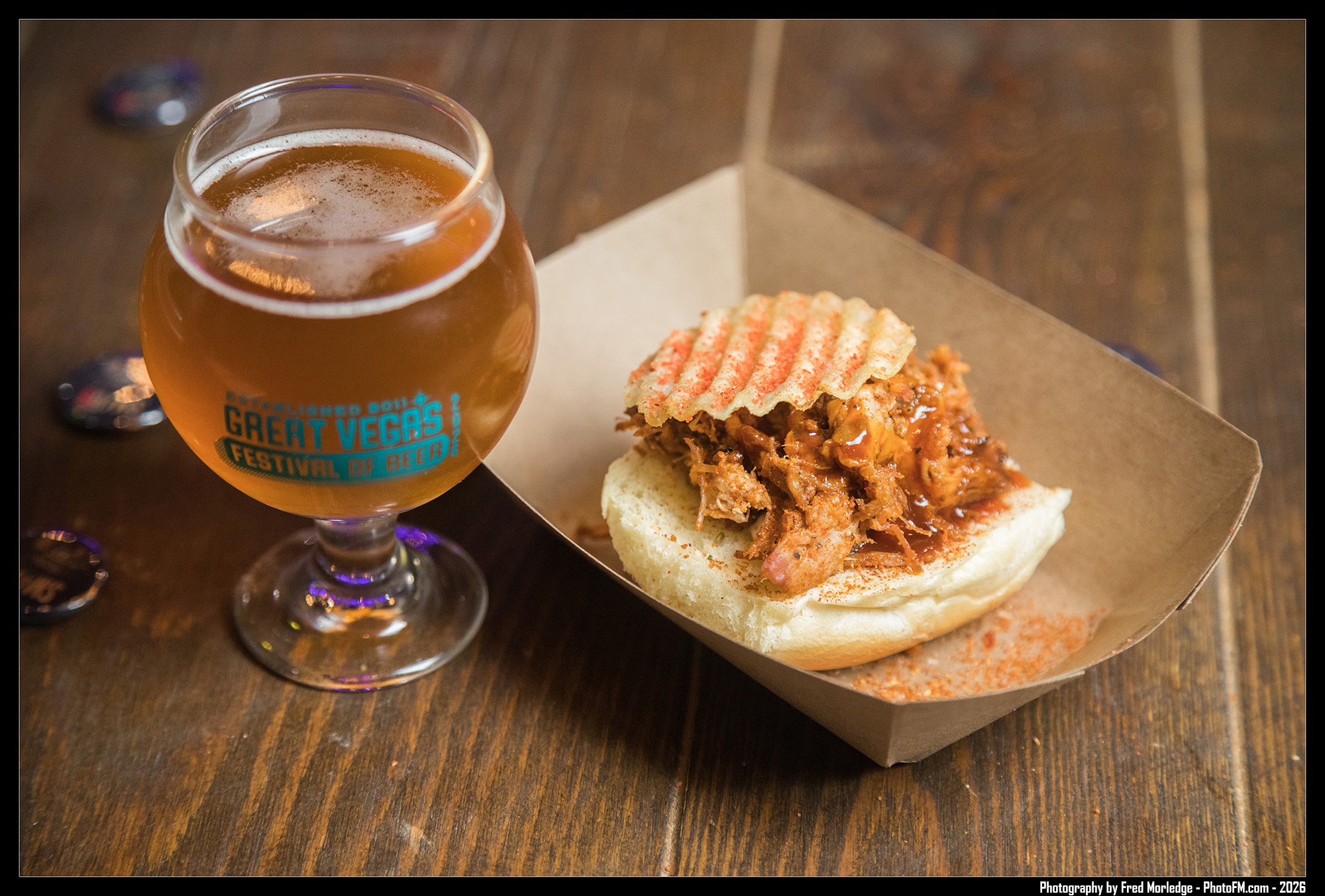 A glass of beer with a logo for the Great Vegas Festival of Beer sitting on a wooden table next to a BBQ sandwich in a cardboard tray with pulled pork and a crispy chip on top.