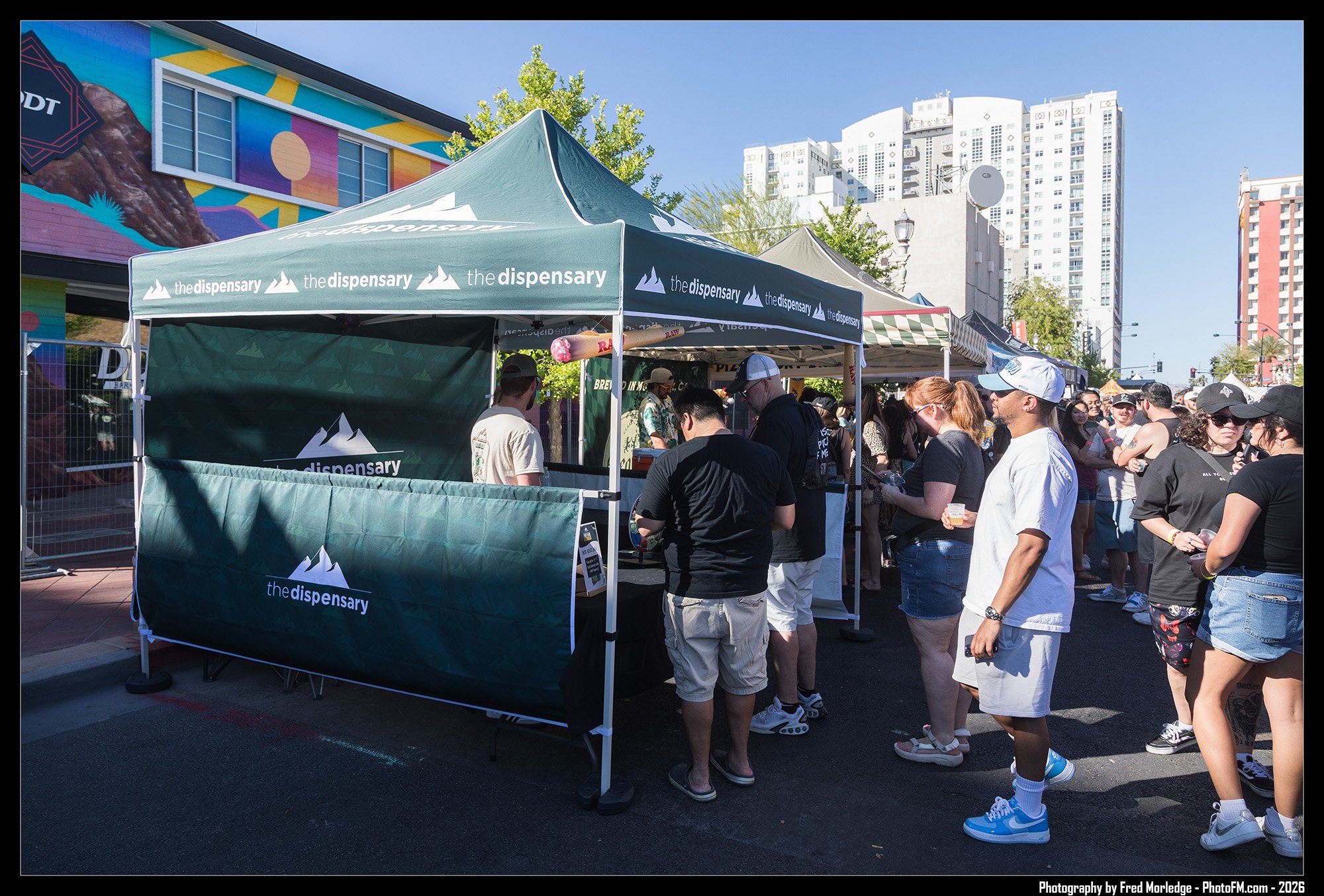 People standing in line at an outdoor vendor booth with a tent that reads 'the dispensary.' Tall buildings and a mural are visible in the background.
