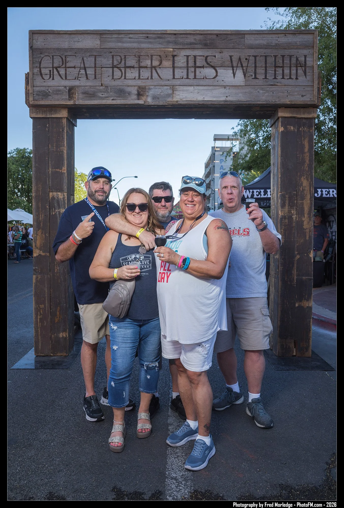 Group of five people posing under a wooden sign that reads 'Great Beer Lies Within' at an outdoor event, with tents and trees in the background.