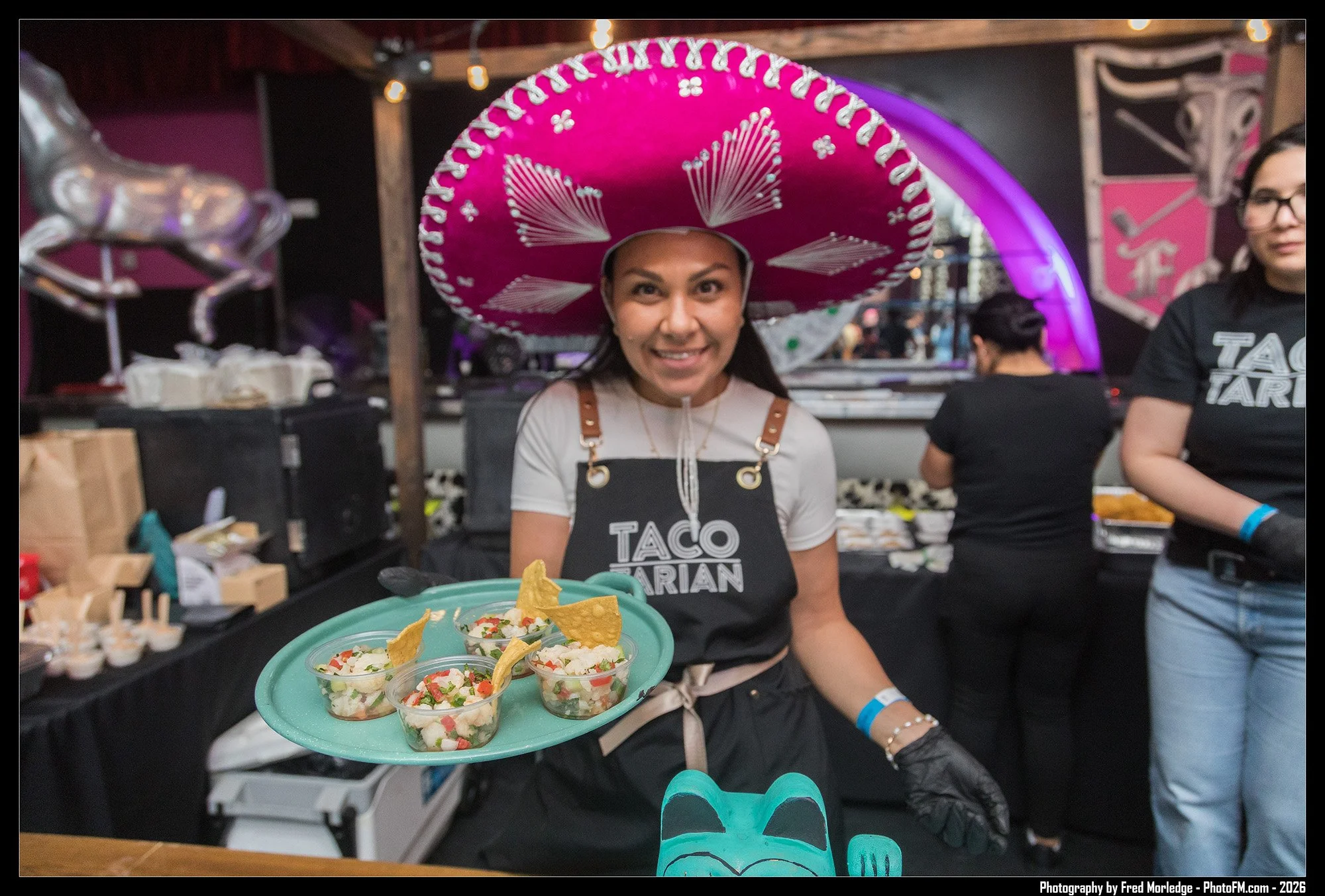 A woman wearing a large pink sombrero hat and a black apron with 'Taco Aria' printed on it, smiling and holding a green tray with cups of chopped ingredients and tortilla chips, in a festive indoor setting with colorful decorations and other people i