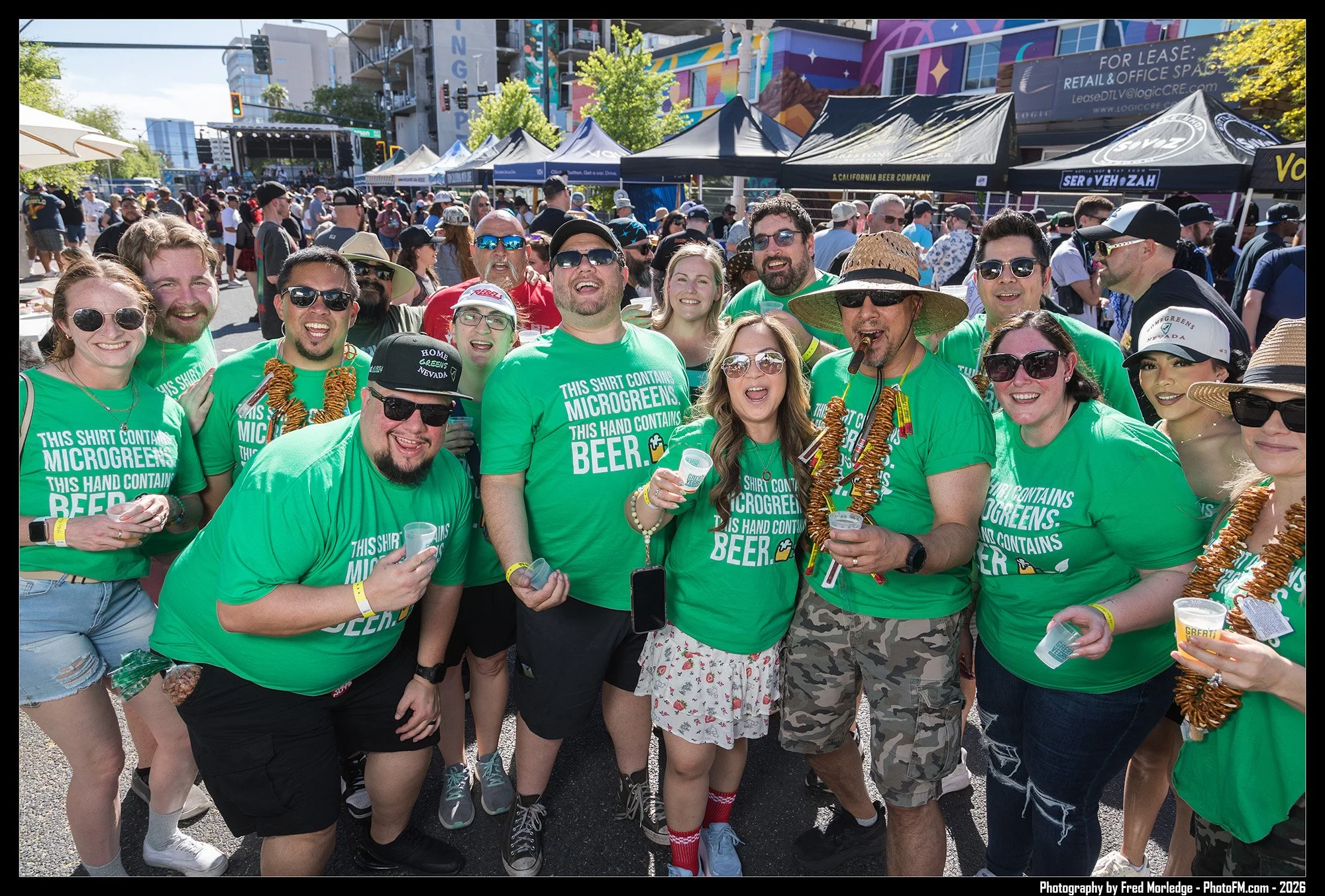 A group of people at an outdoor event celebrating, many wearing matching green shirts with text about Microgreens and beer, some with leis and hats, holding drinks, with food stalls and a stage in the background.