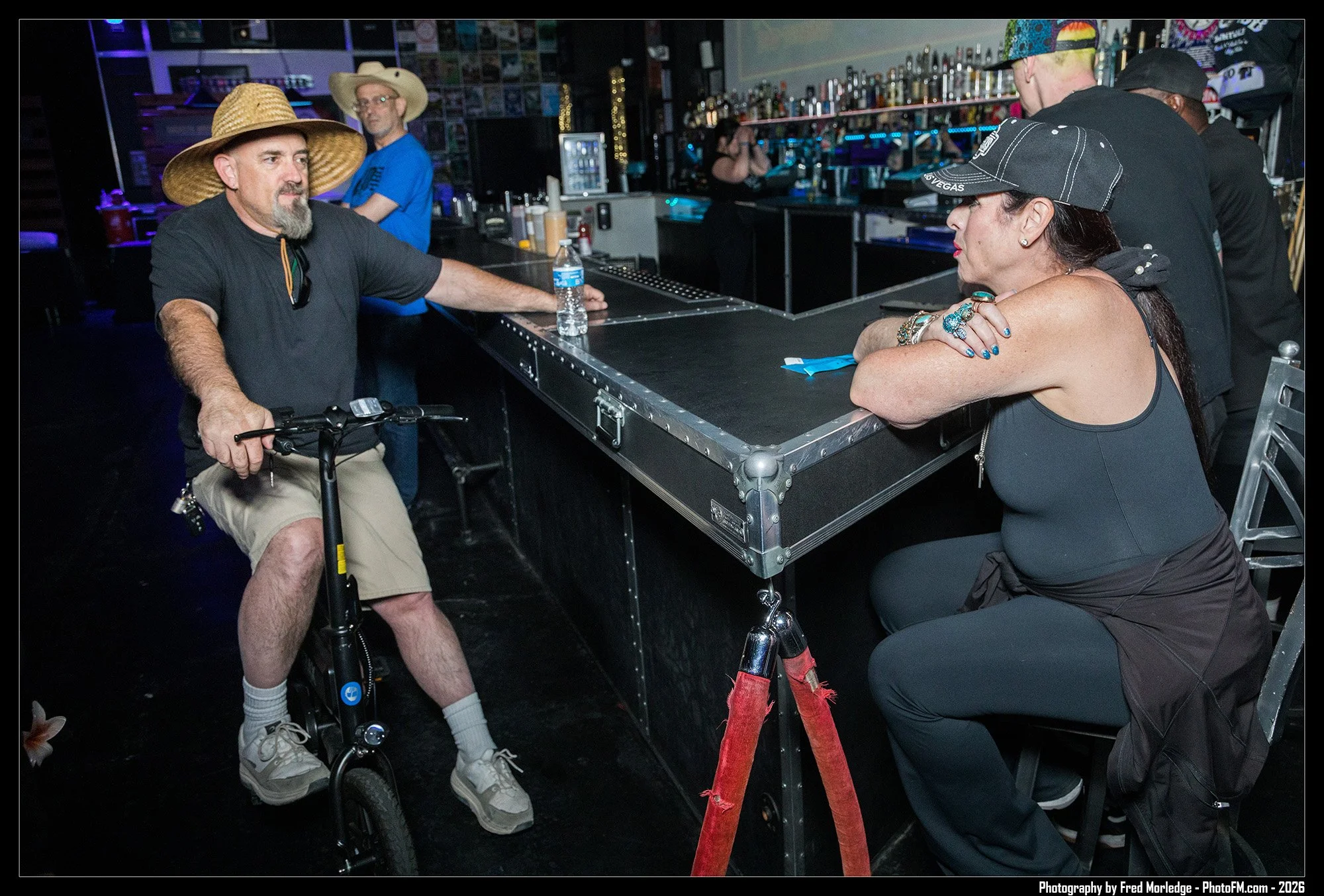 A man in a black shirt and tan shorts sitting on a mobility scooter at a bar counter, wearing a large straw hat. He is talking to a woman with dark hair, wearing a baseball cap, with her arms on the bar. In the background, there are bar shelves with 