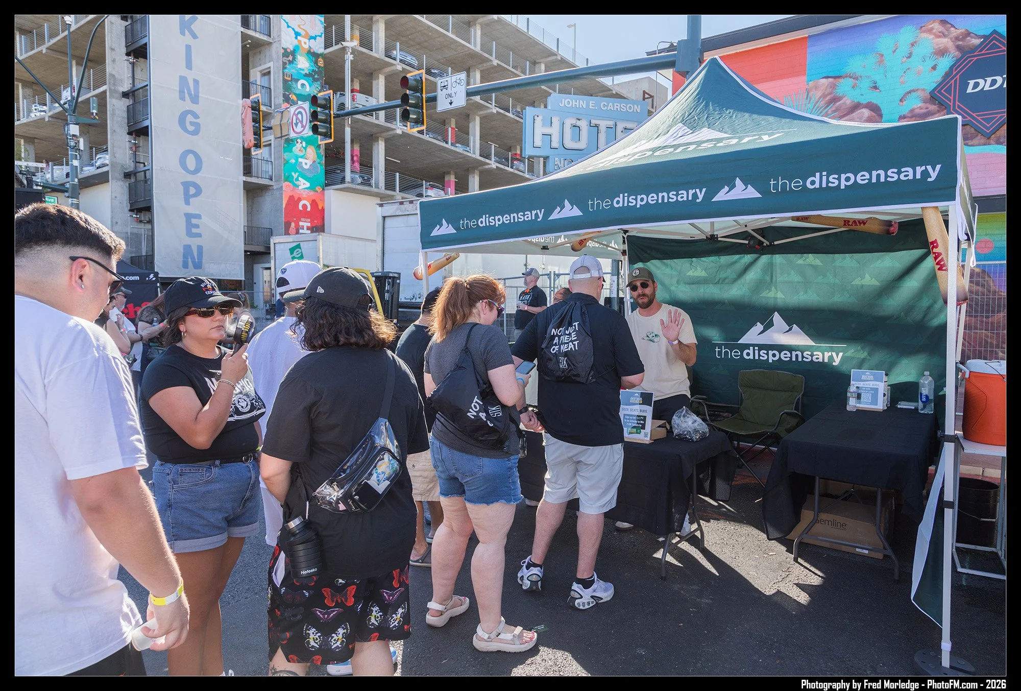 People standing in line at an outdoor cannabis dispensary booth called 'the dispensary' with a green canopy, in an urban setting with a parking garage and colorful mural in the background.