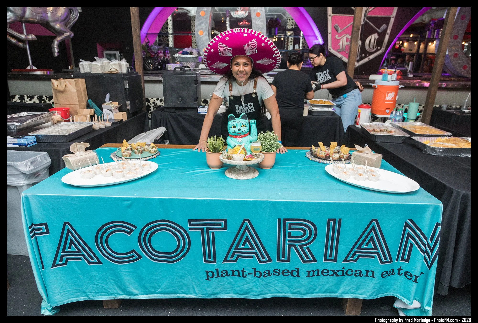 A woman wearing a pink sombrero stands behind a table with a blue tablecloth that says 'Plant-based Mexican eater'. The table displays desserts, drinks, and decorations. In the background, two women prepare food at a taco stand, and there are decorat