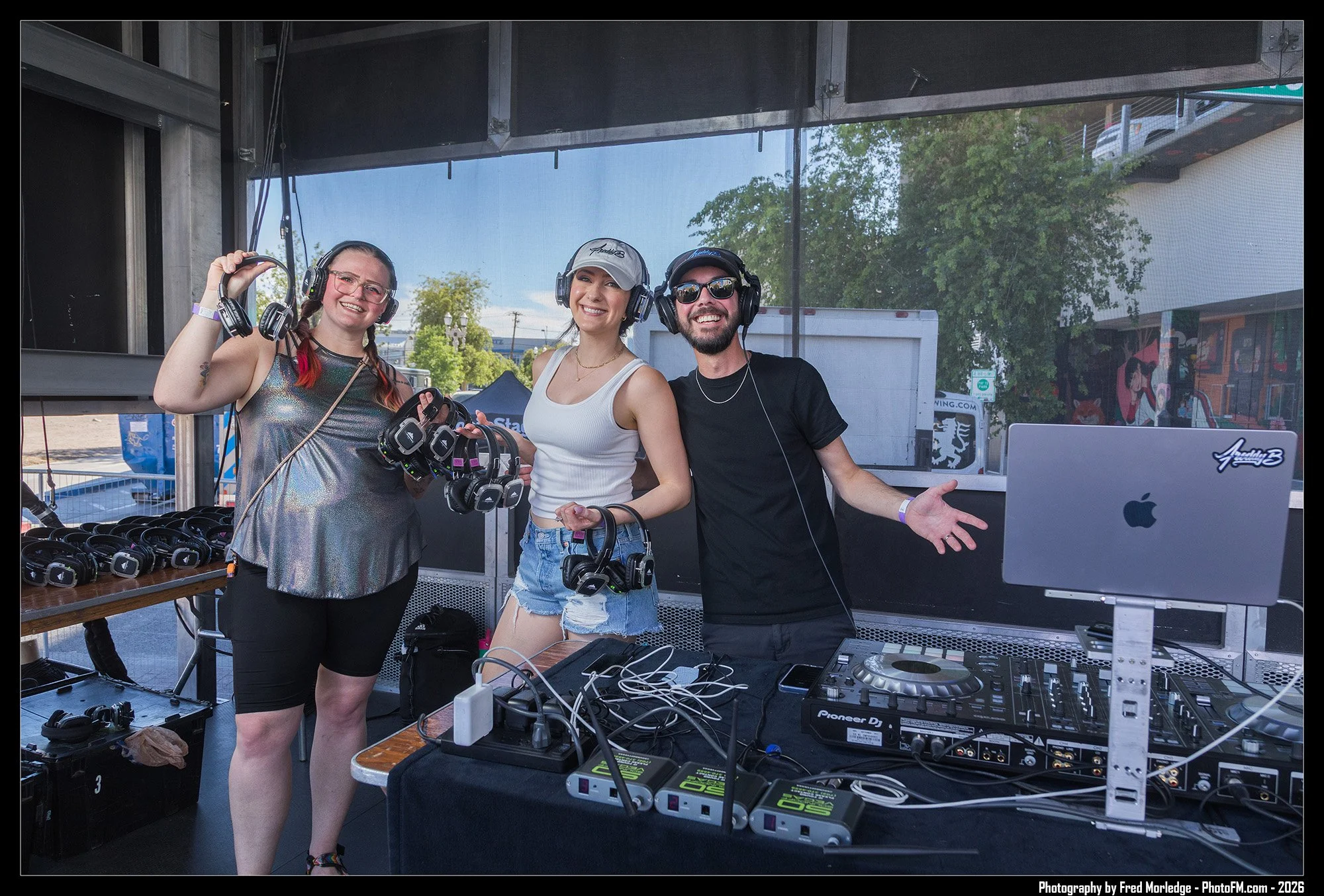Three young people with headphones and DJ equipment at an outdoor event, smiling and posing for the photo.