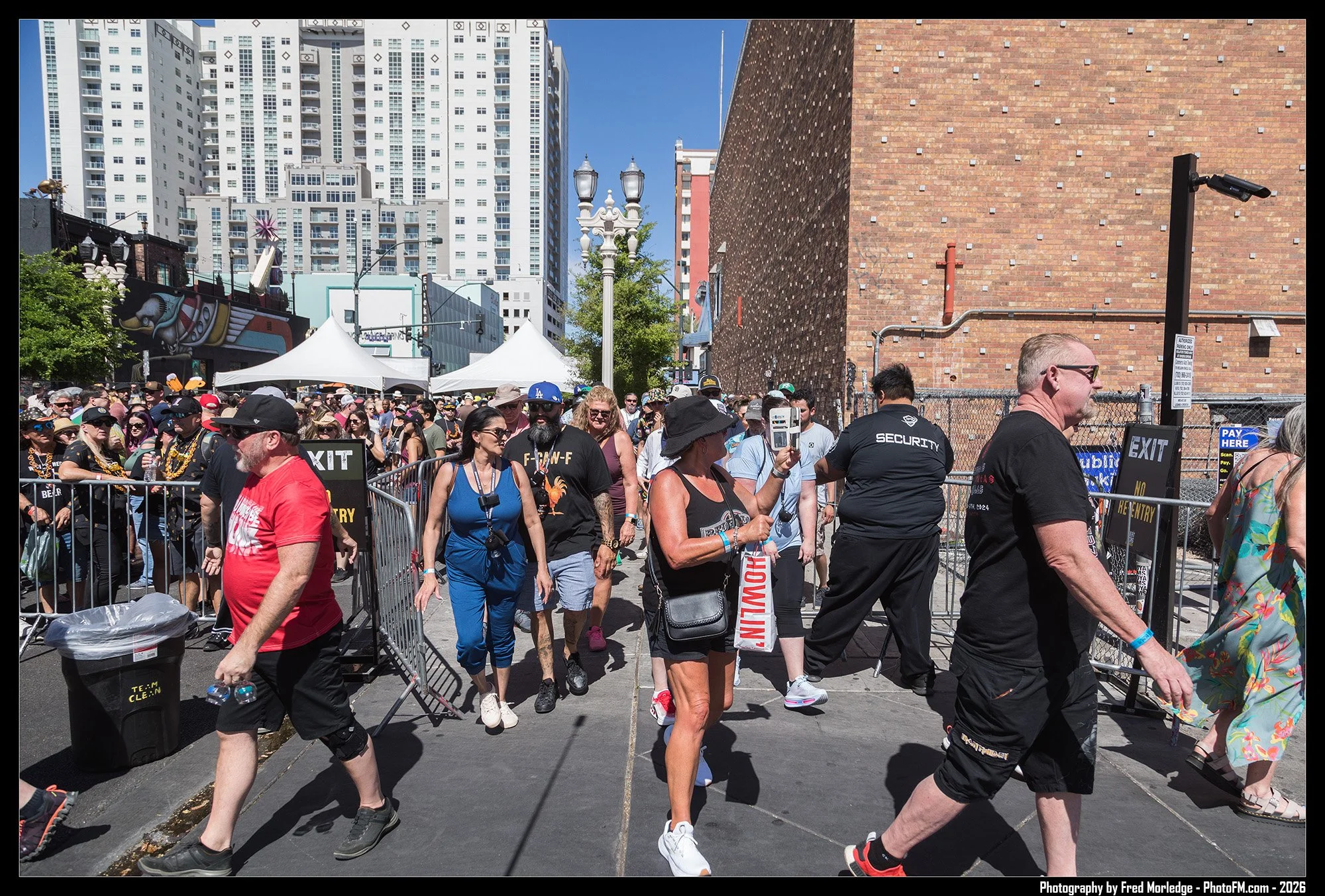 Crowd of people walking near a fenced area on a city street during daytime, with high-rise buildings in the background and tents set up for an event.