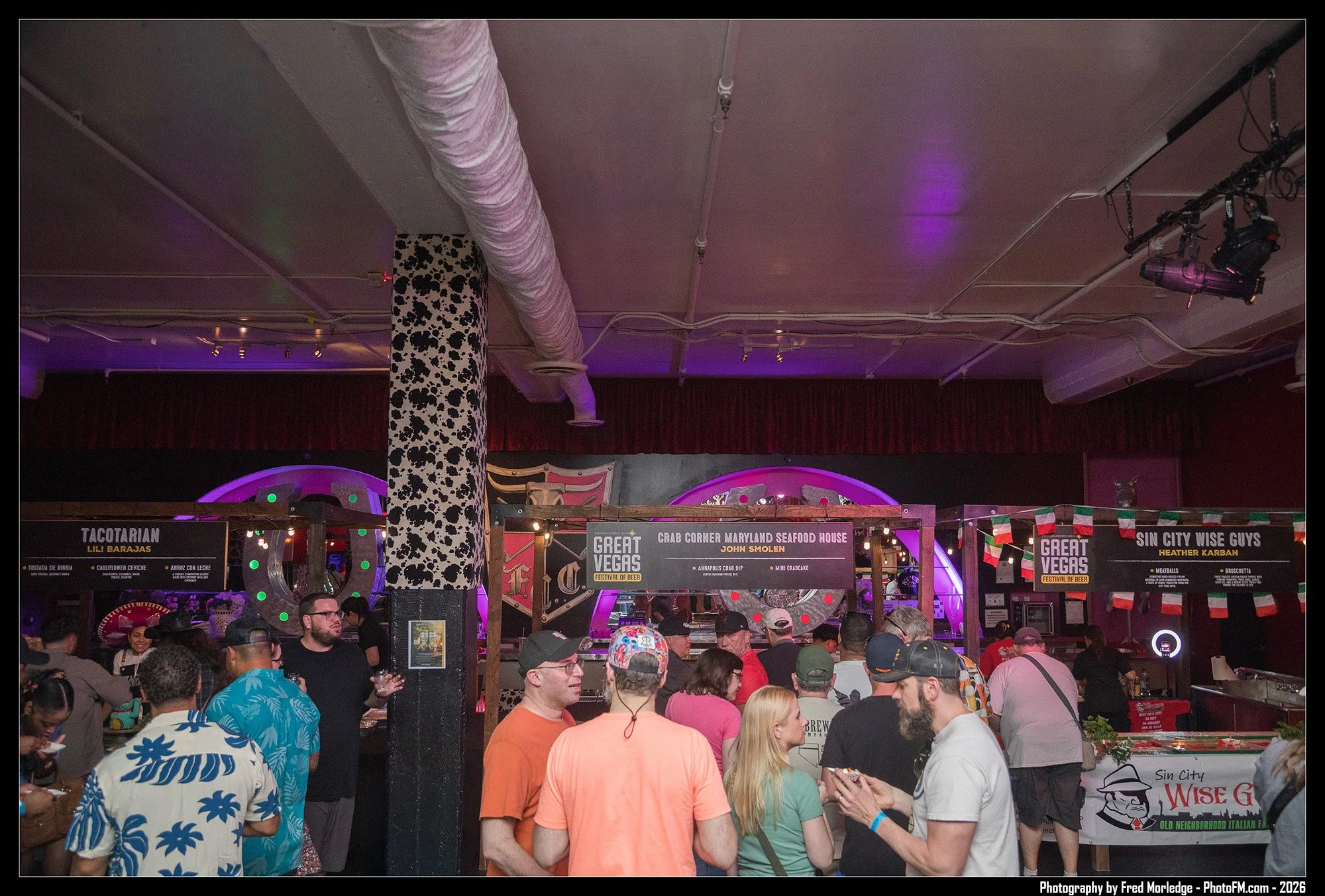 Crowd of people waiting in line at a food festival with various food stalls and banners, illuminated by purple lights, indoor setting.
