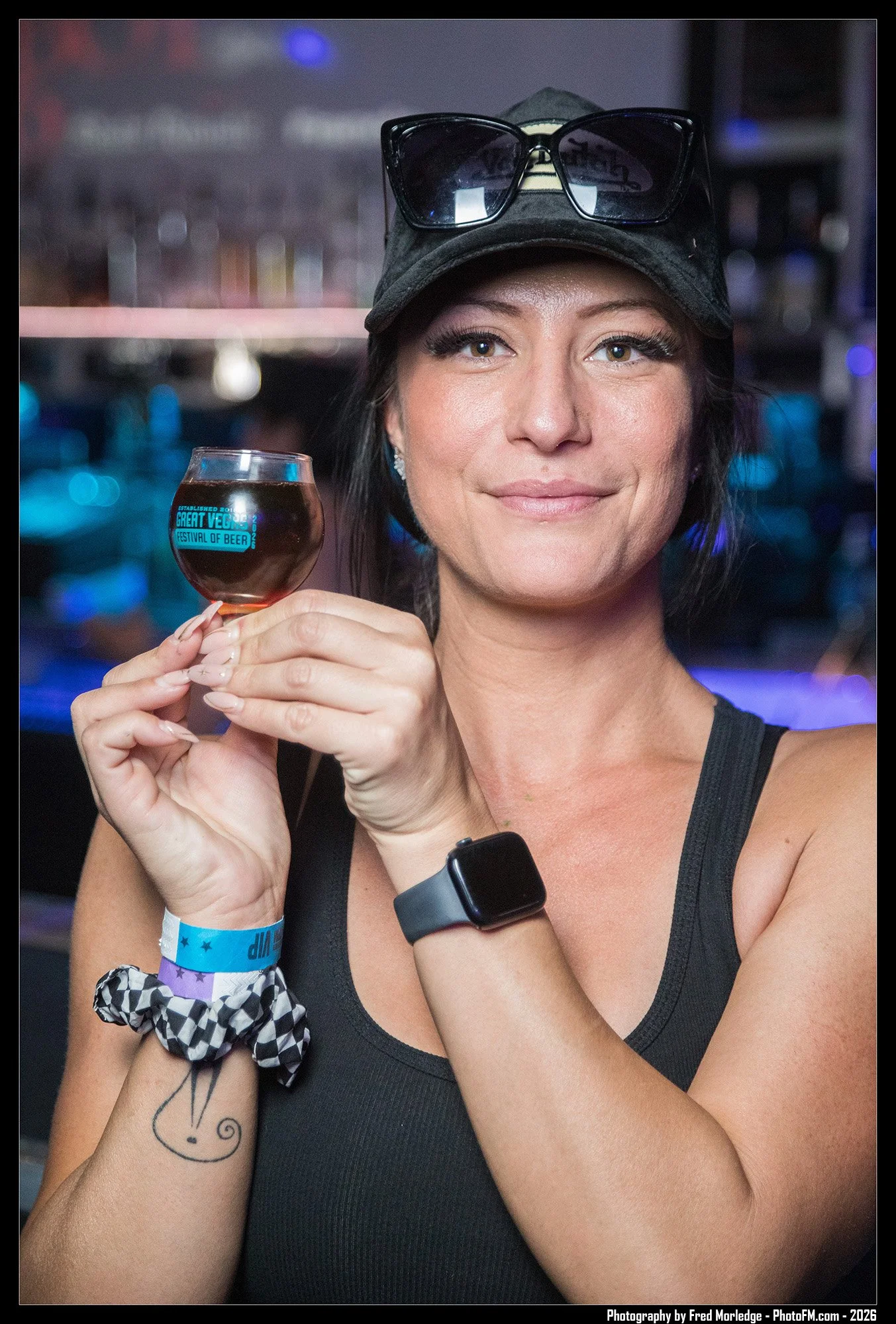 A woman with a light complexion, wearing a black tank top, a black cap with sunglasses on it, holding a glass of beer. She has long eyelashes and a tattoo of a simple line drawing of a face on her forearm, along with a black and white checkered scrun
