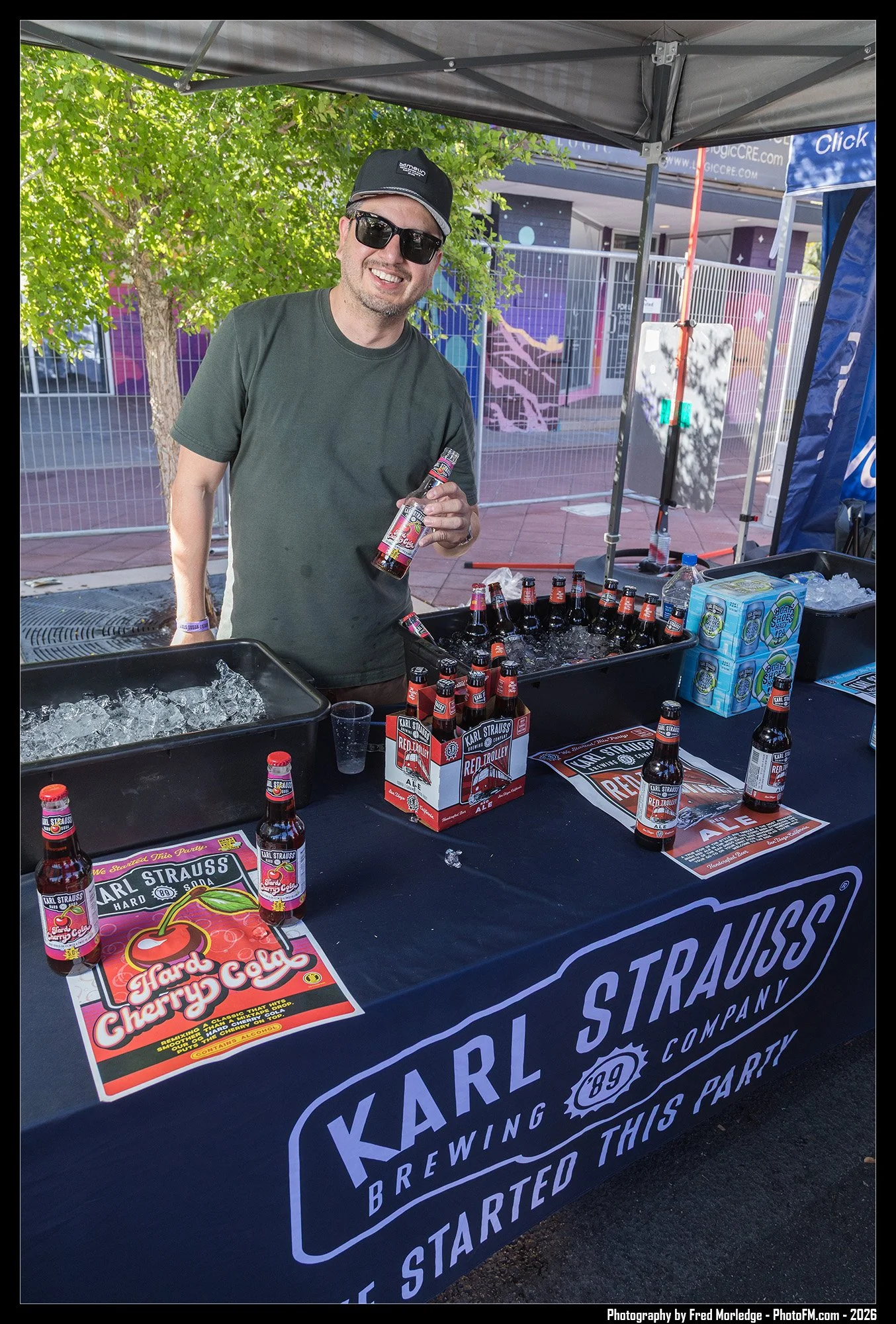 A man smiling at a stand promoting Karl Strauss Brewing Company Cherry Cola, holding a bottle of flavored soda. The stand has bottles of Cherry Cola, advertising posters, and a black tablecloth with the company's logo.