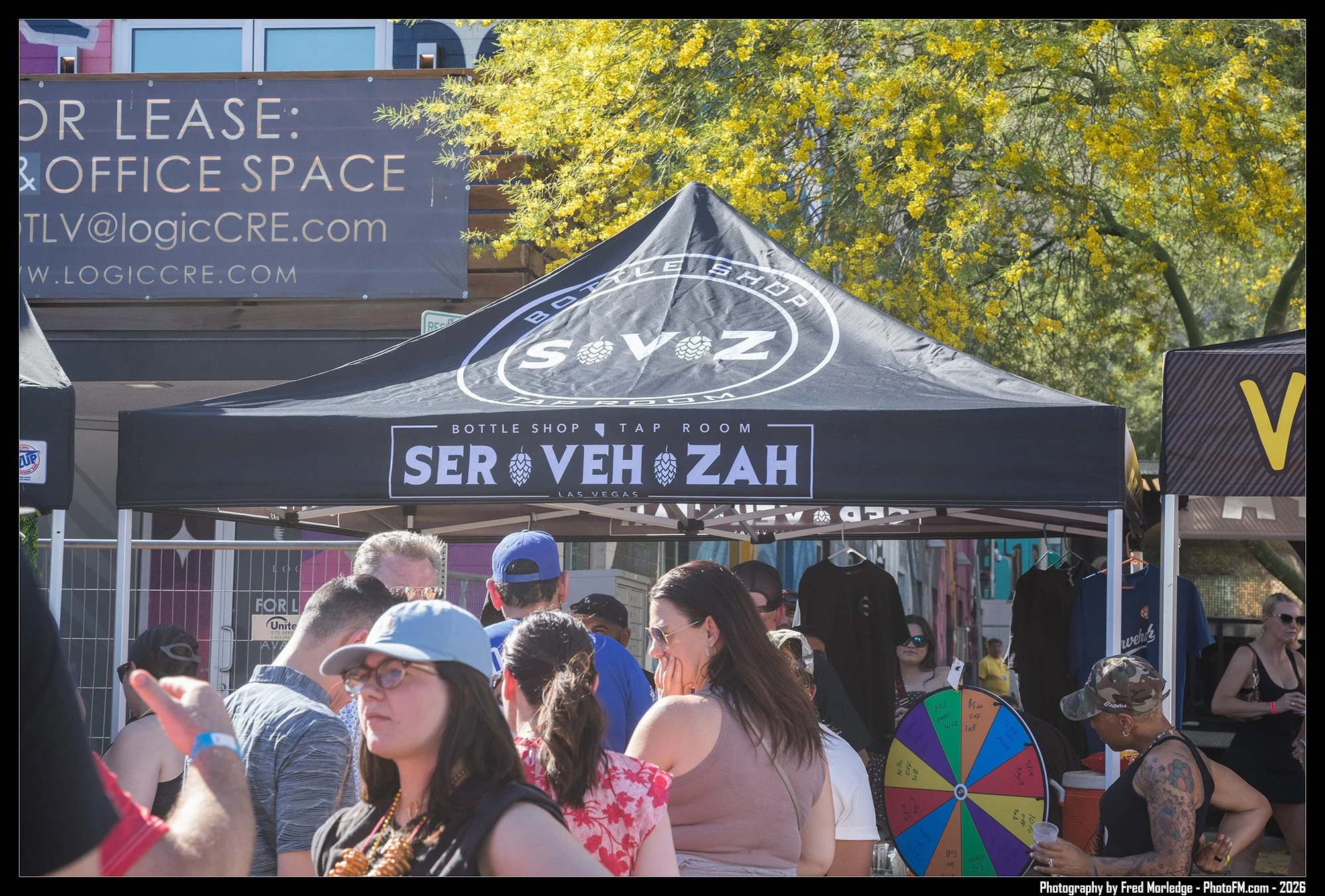 A crowd of people gathered at an outdoor event with a black canopy that reads 'Bottle Shop' and 'Ser Ve Zah' in Vegas. There is a colorful spinning wheel behind them, and some people are wearing sunglasses and hats. In the background, there are trees