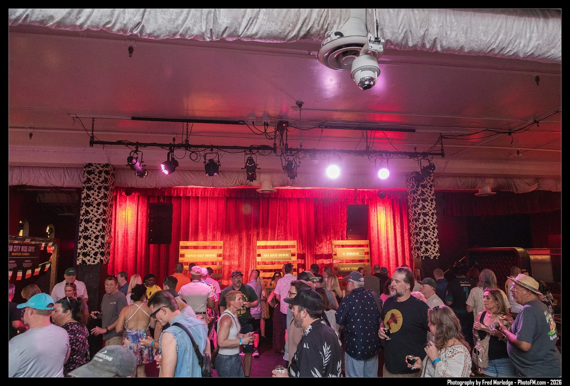 Crowd of people attending an indoor event with a stage featuring red curtains, illuminated lights, and signs indicating breweries.