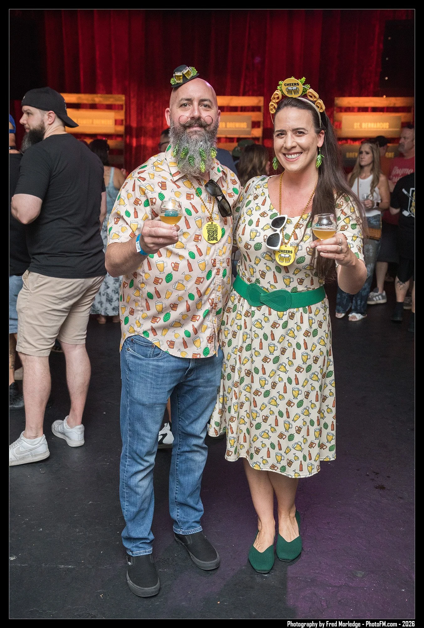 A man and woman dressed in matching festival outfits with beer-themed patterns, holding small glasses of beer, at a lively indoor event with a red curtain backdrop and other people in the background.