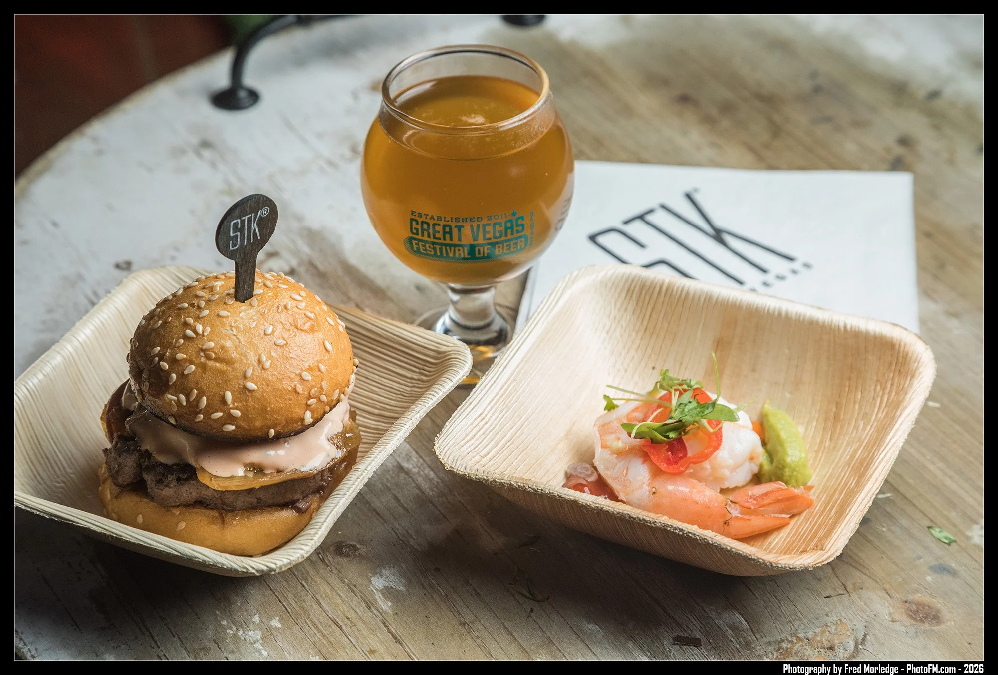 A burger with sesame seed bun, beef patty, cheese, pickles, and sauce in a disposable container. Next to it, a seafood dish with shrimp, vegetables, and sauce in a separate container. In the background, a glass of beer with a logo from the Great Vega