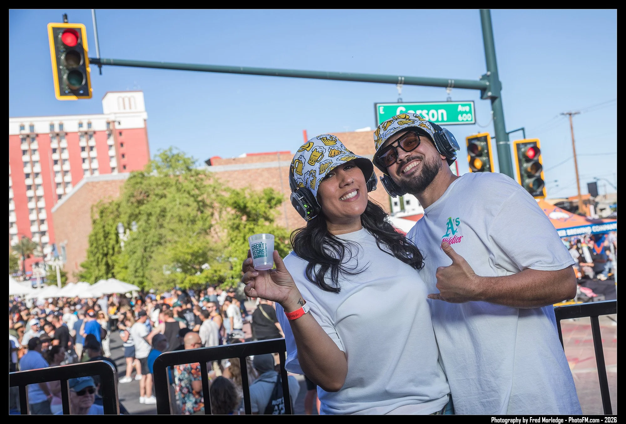 Two smiling people, a woman and a man, wearing headphones and baseball caps with beer and crown designs, standing in front of a crowd at an outdoor event. The woman is holding a cup that says "Great Vegas". There are traffic lights and a street sign 