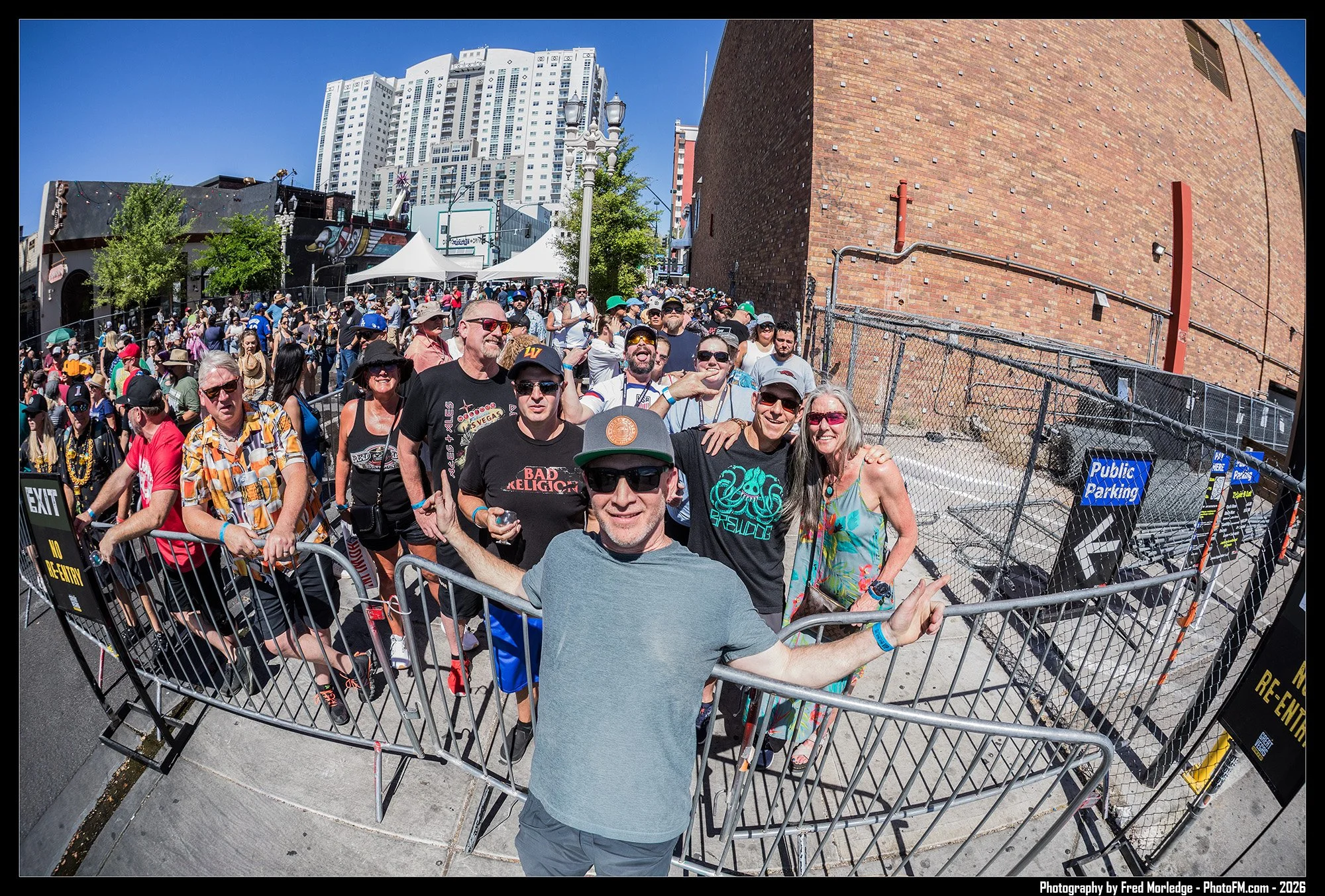 A group of smiling people standing behind portable fencing outside on a sunny day, with a tall city building and brick wall in the background.