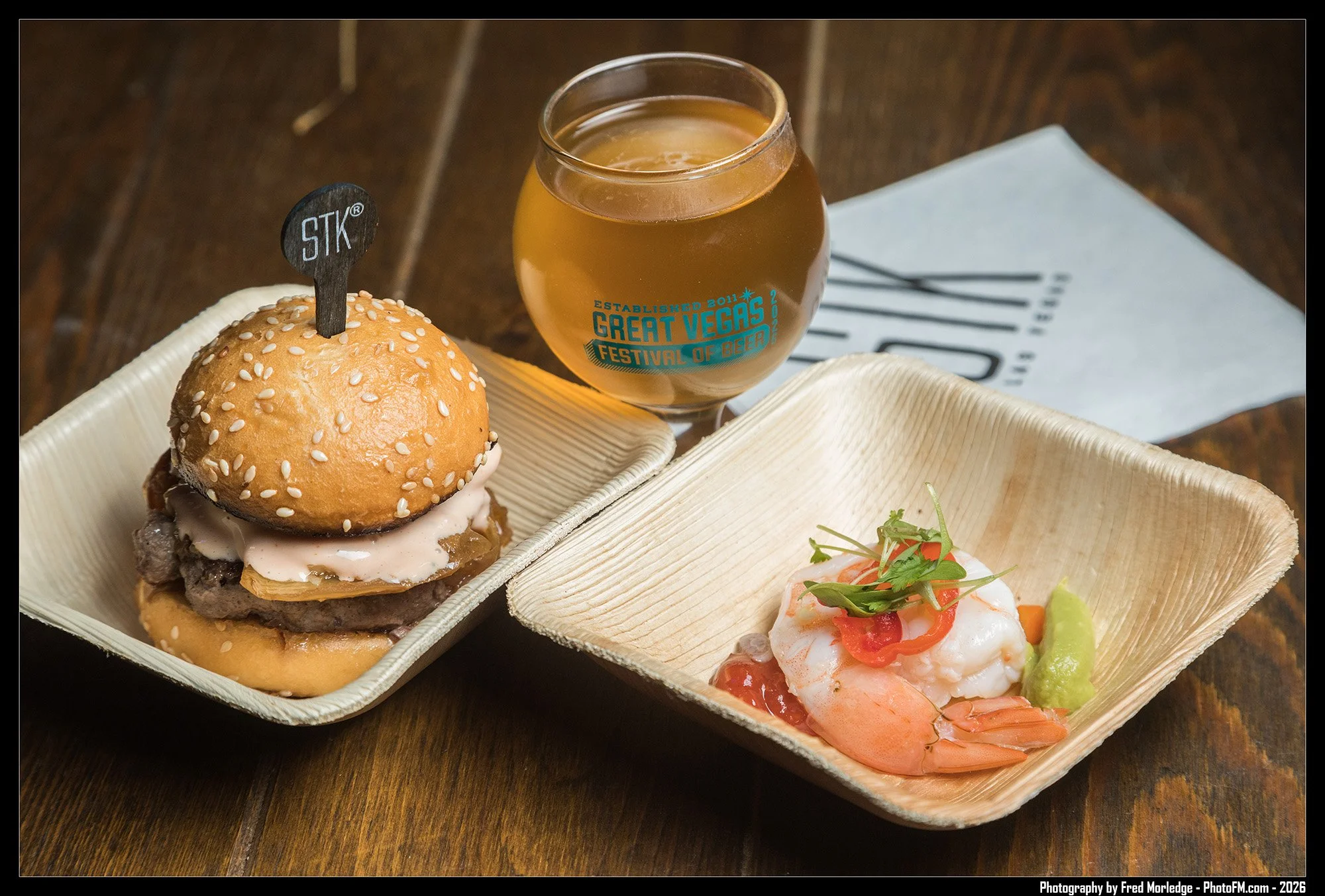 A tray with a burger topped with sesame seed bun, lettuce, onion, cheese, and burger patty, a glass of beer with a logo, and a small plate with shrimp, cherry tomatoes, cilantro, lime, and sliced vegetables.