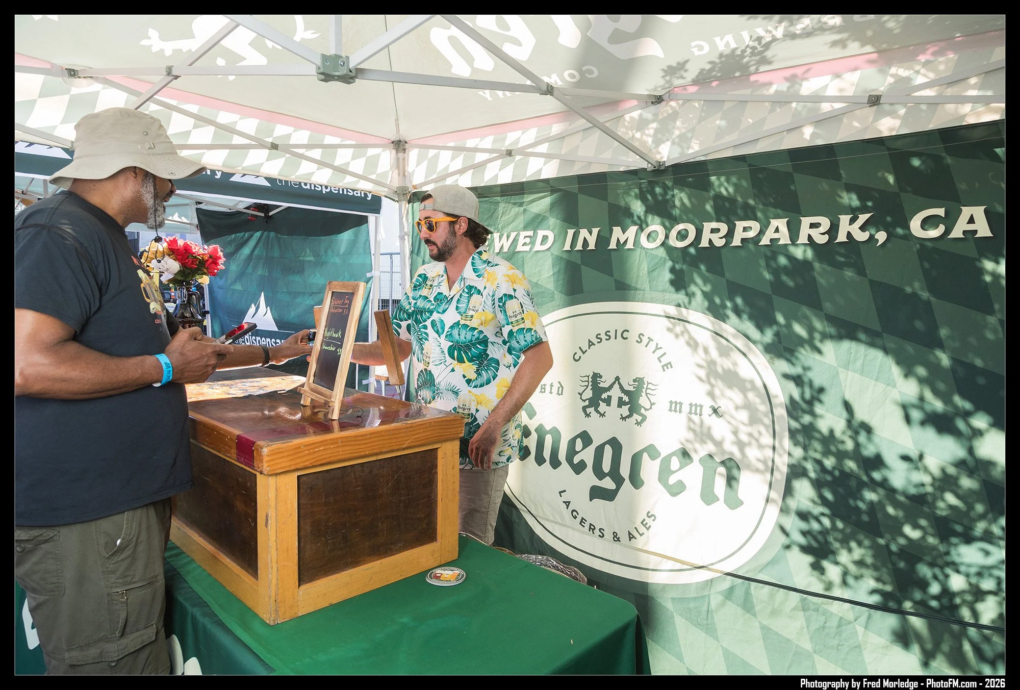 A man in a gray baseball cap and tropical shirt standing at a booth with a wooden crate on a green table, talking to another man wearing a hat and glasses. The booth has a green banner with white text, indicating it is in Moorpark, California, and ad