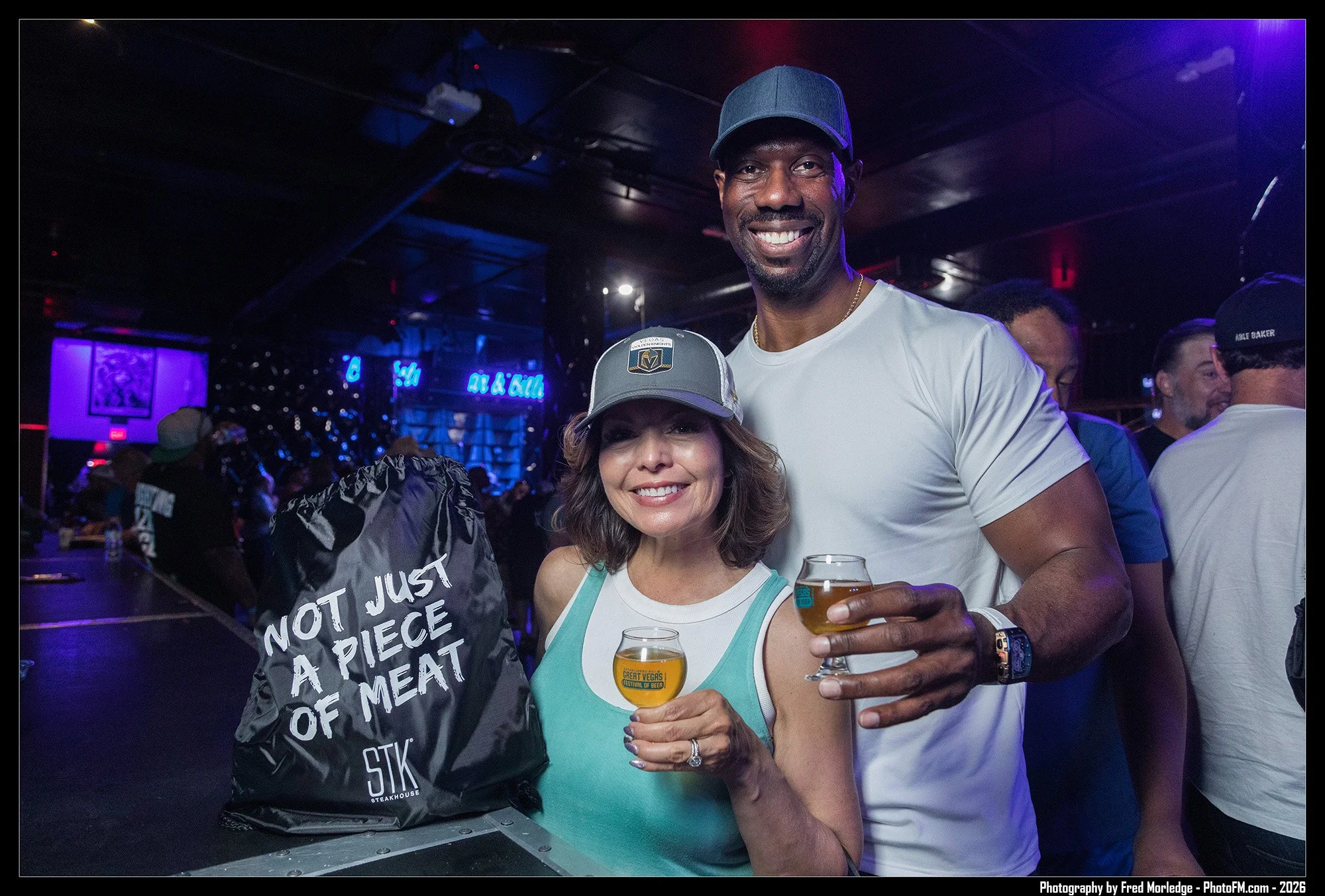 A smiling man and woman holding glasses of beer at a bar or brewery, with people and neon lights in the background.