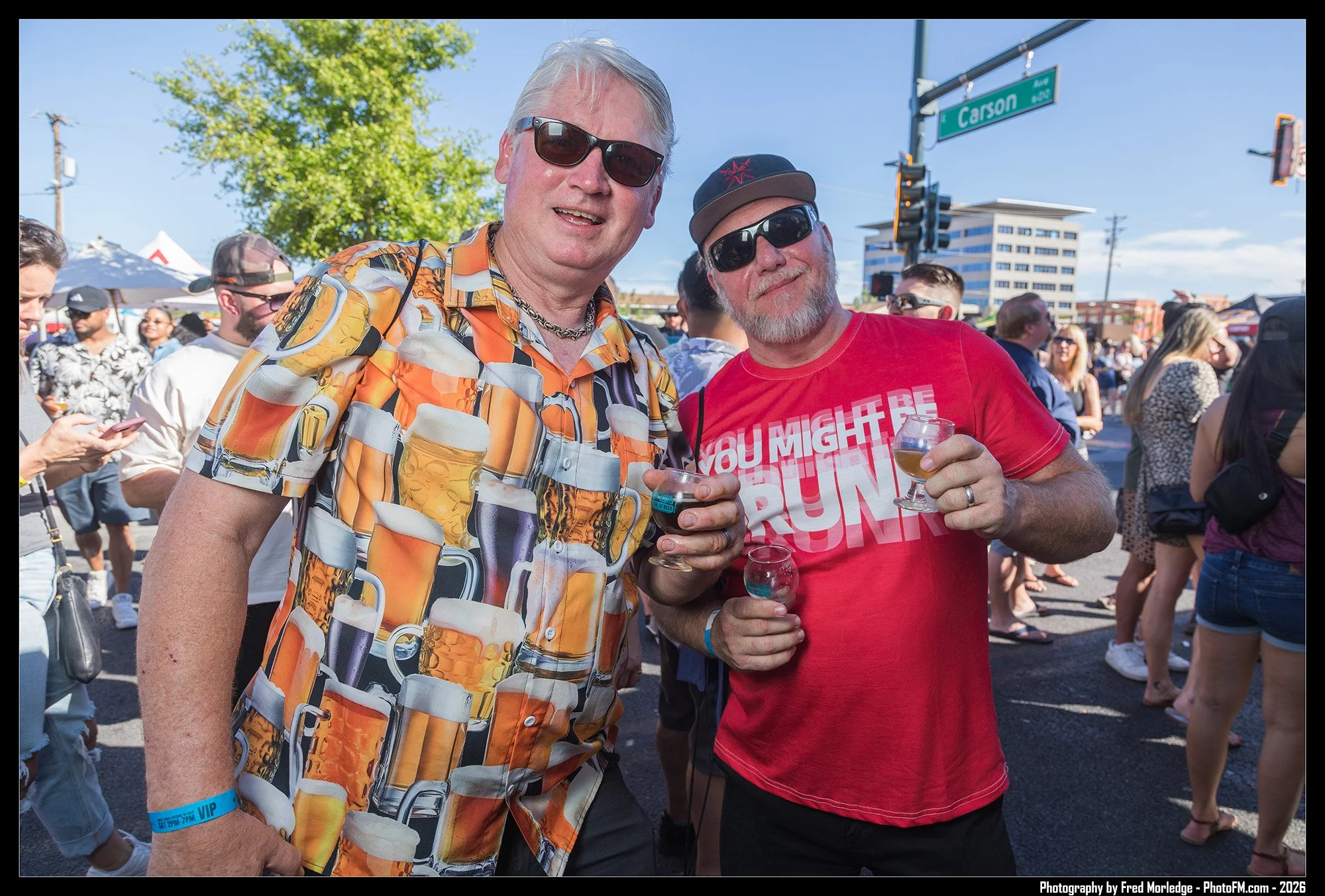Two men standing together at an outdoor event, holding glasses of beer and wearing sunglasses, amidst a crowd of people.