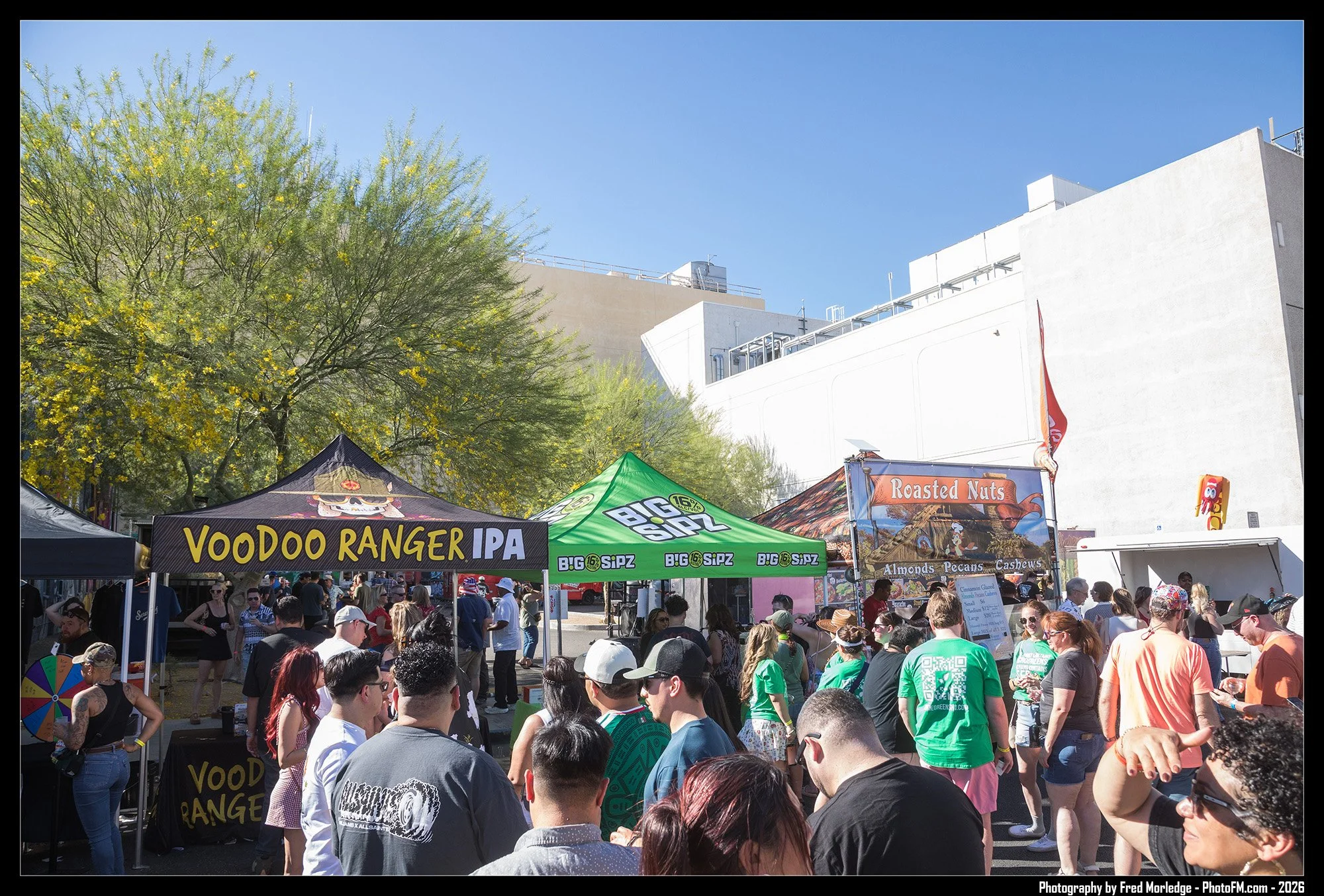 Crowd of people at an outdoor festival or market with vendor tents, including one labeled Voodoo Ranger IPA and another offering roasted nuts and almonds, cashews, and pecans, under blue skies with trees and buildings in the background.