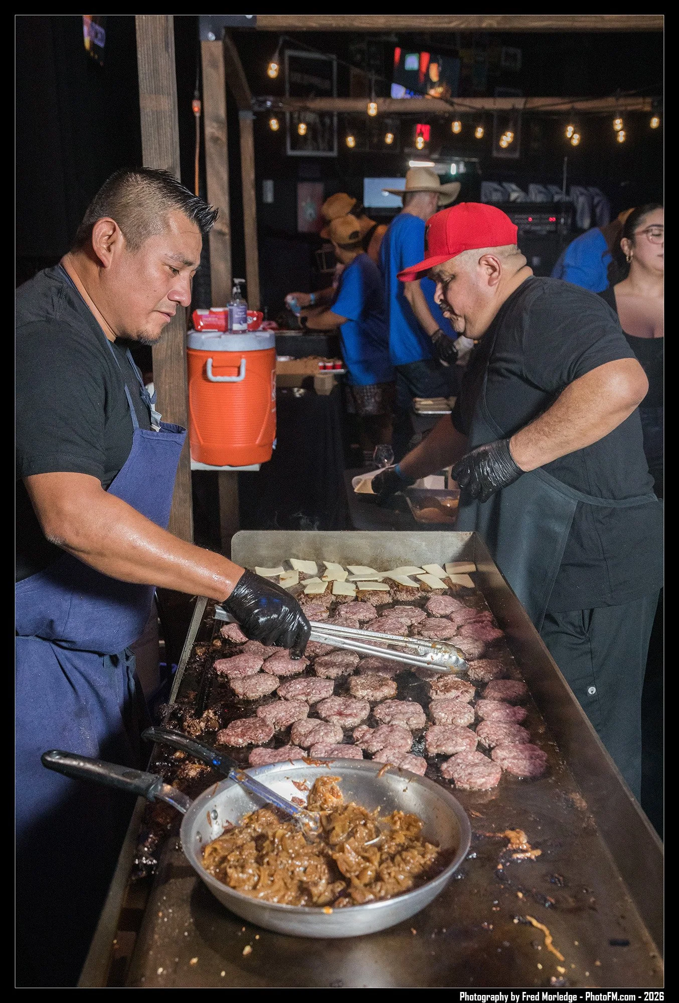 Two cooks are preparing burgers on a grill at a restaurant or food stall. One chef is using tongs to handle burger patties, while a tray of cooked meat and a bowl of seasoned ingredients are on the counter. In the background, there are people, some w