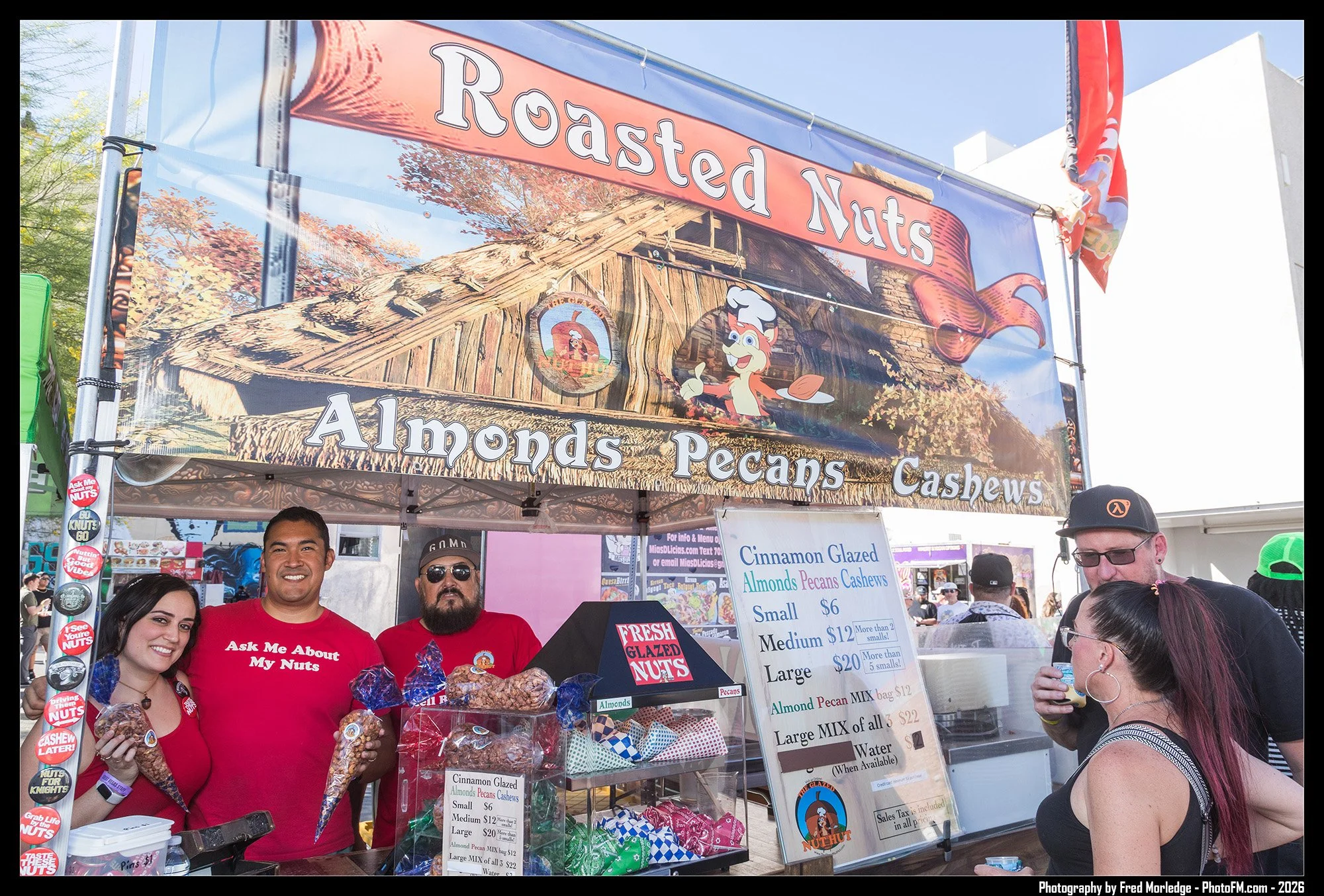 A street fair or festival with a booth selling roasted nuts, almonds, pecans, and cashews. Four people are posing and smiling behind the booth, one wearing a red shirt with 'Ask Me About My Nuts' written on it. A children or cartoon character mascot 