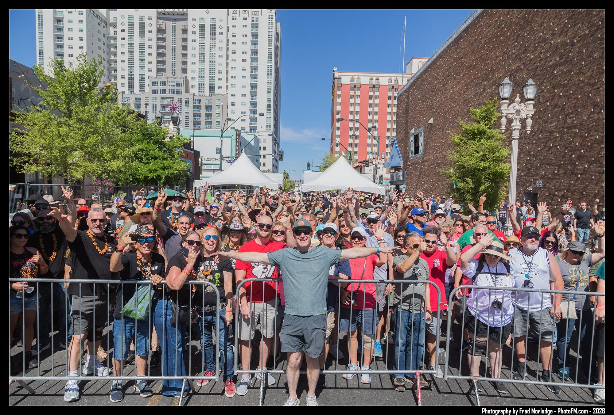 Crowd of people standing behind a metal barricade at an outdoor event on a sunny day with tall buildings in the background.