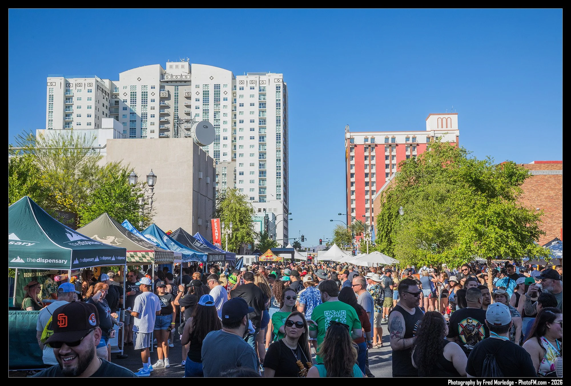 A bustling outdoor street festival with many people walking and standing under tents, against a backdrop of tall buildings and clear blue sky.
